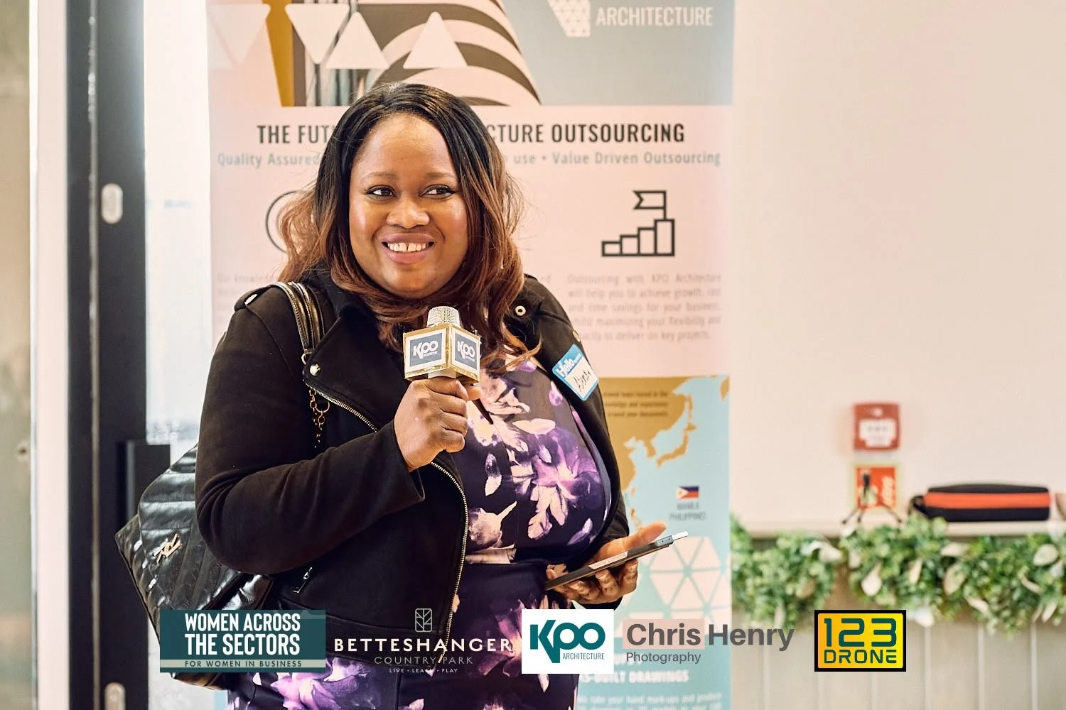 A woman holding a microphone and a tablet, smiling at an indoor event with banners and greenery in the background.