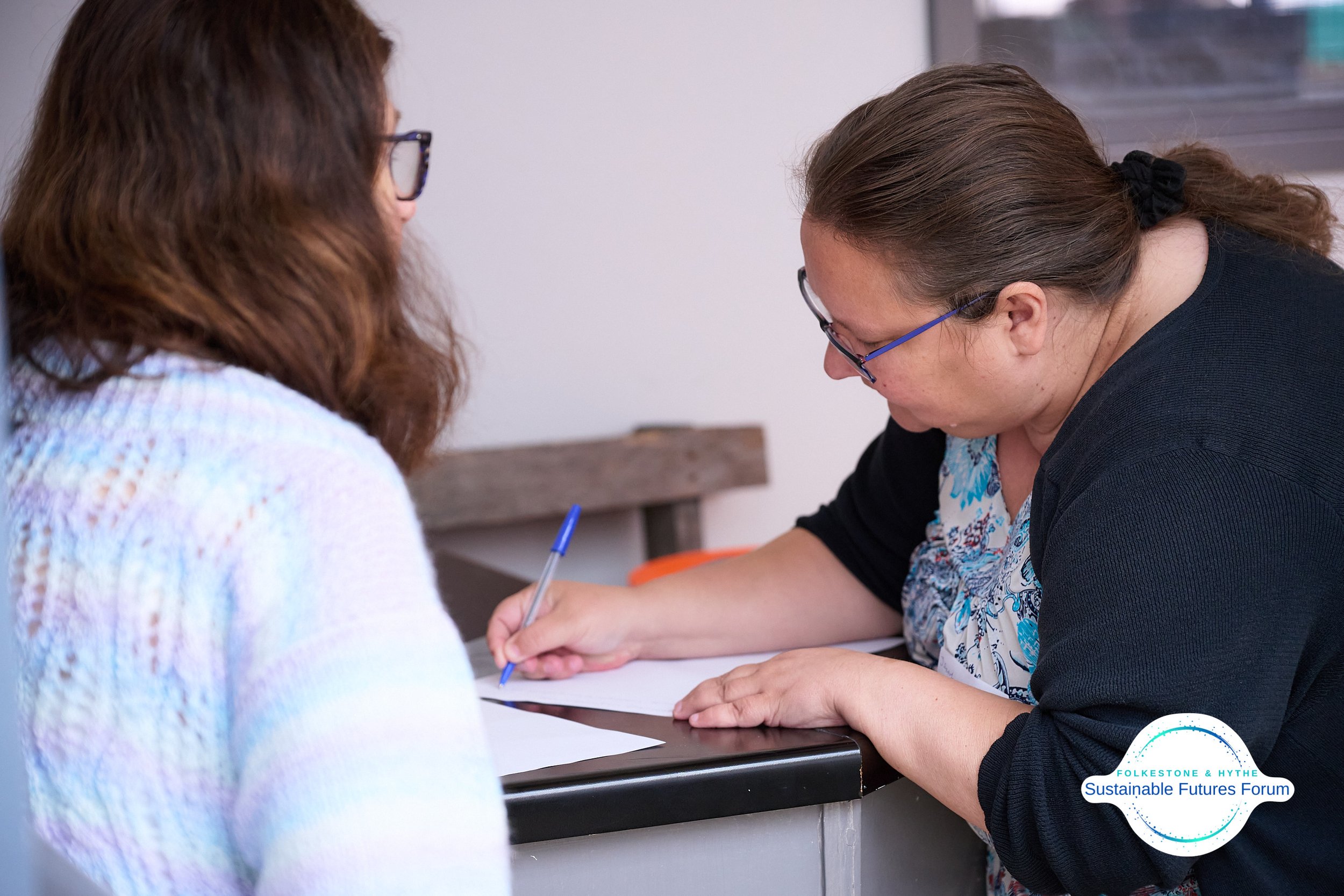 Two women sitting at a desk, one writing on paper, engaged in conversation at a Sustainable Futures Forum event.
