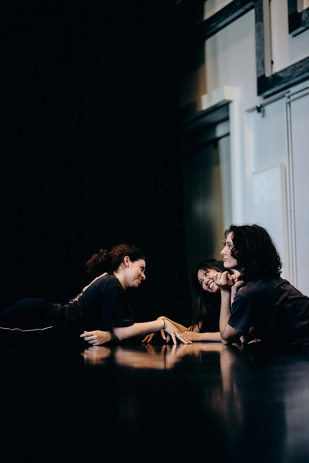 Three women sit at a dark table, smiling and engaging in conversation, with a dark background and a bright window behind them.