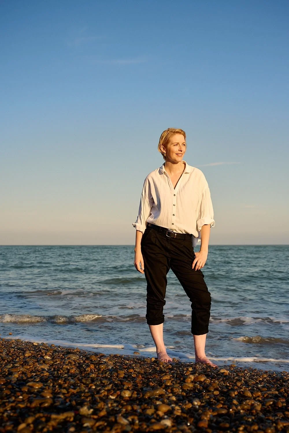 A woman standing barefoot on a pebble beach with the ocean and a clear blue sky in the background.