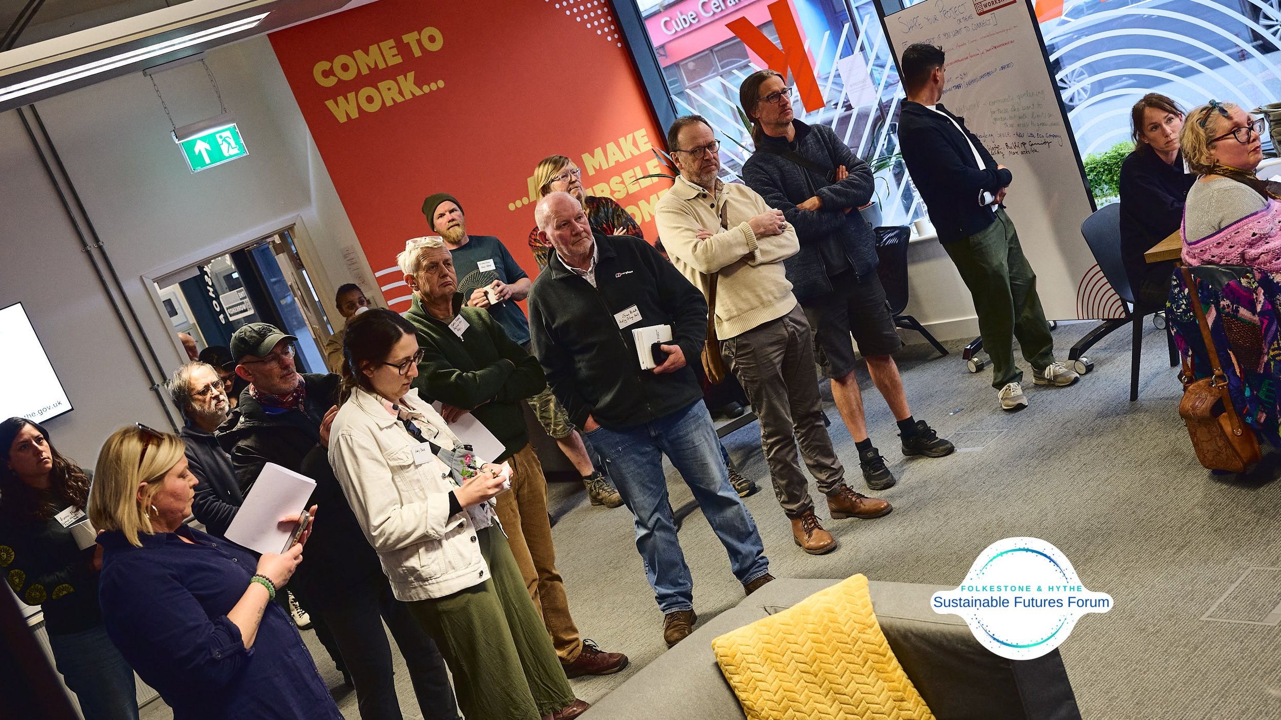 A group of people attending a presentation or discussion at the Folkestone & Hythe Sustainable Futures Forum, standing and seated, in a modern room with a red wall, large windows, and a whiteboard, some taking notes and others listening attentively.