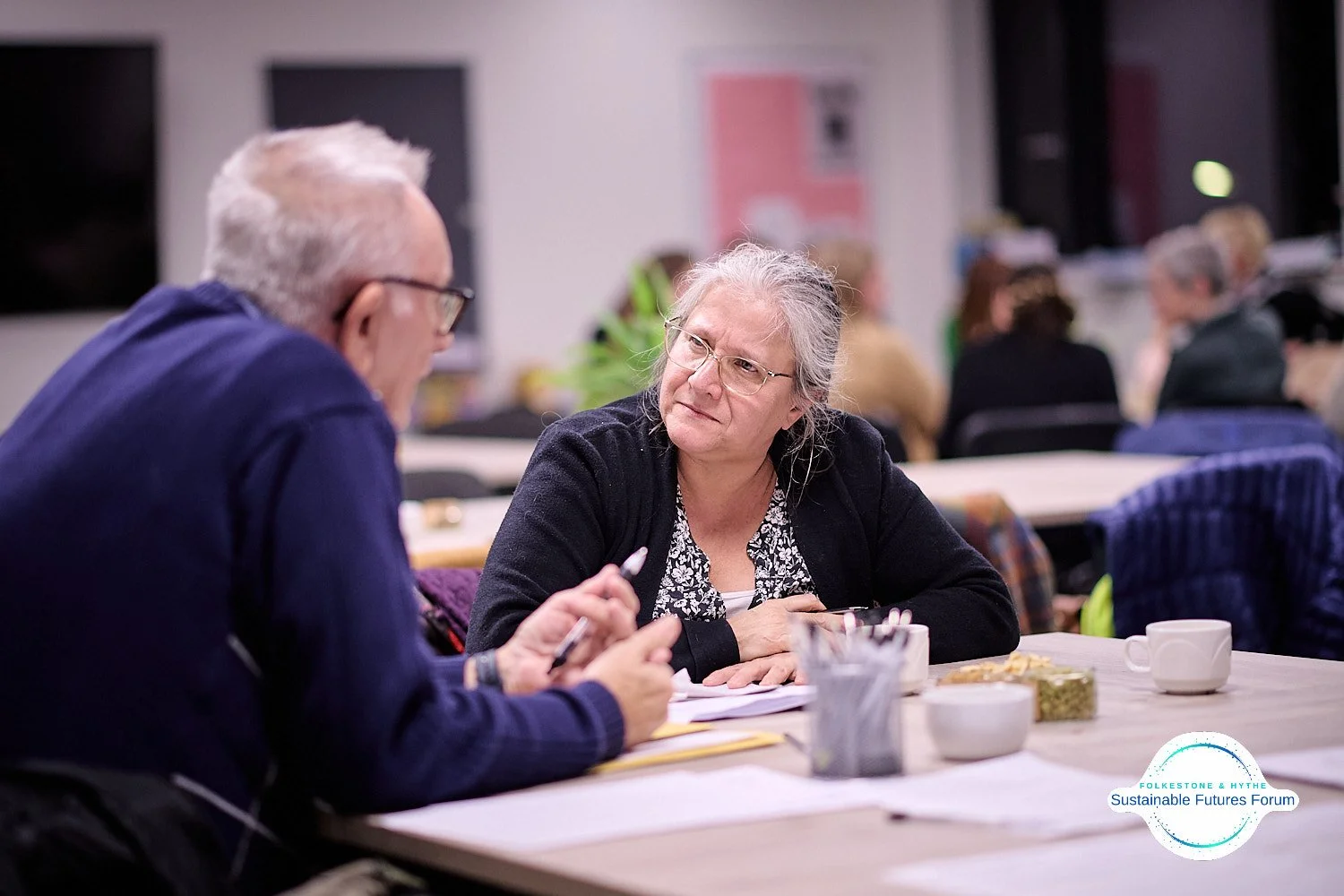 An older woman with gray hair and glasses engaged in conversation with a man at a table during a forum event, with other attendees in the background.