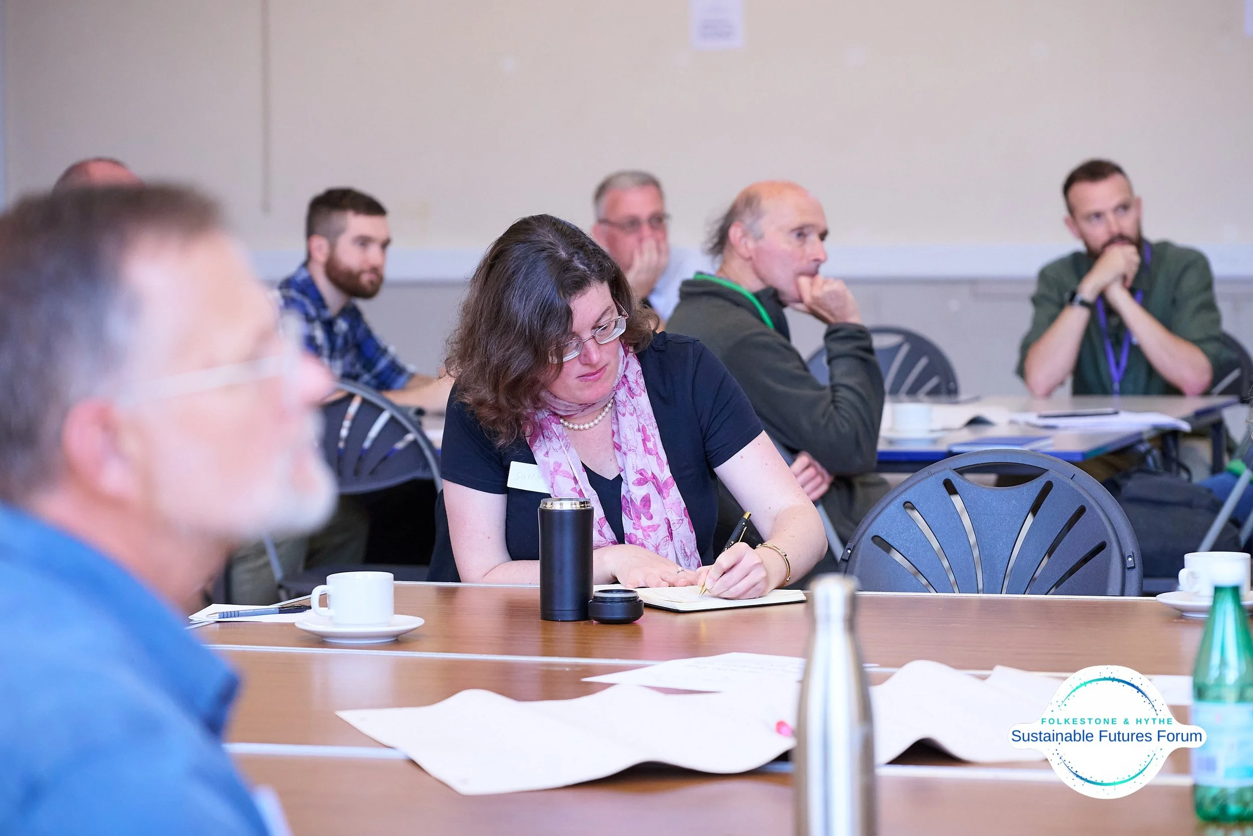 People attending a workshop or conference around tables, taking notes, with cups and papers, in a room labeled 'Folkestone & Hythe Sustainable Futures Forum'.