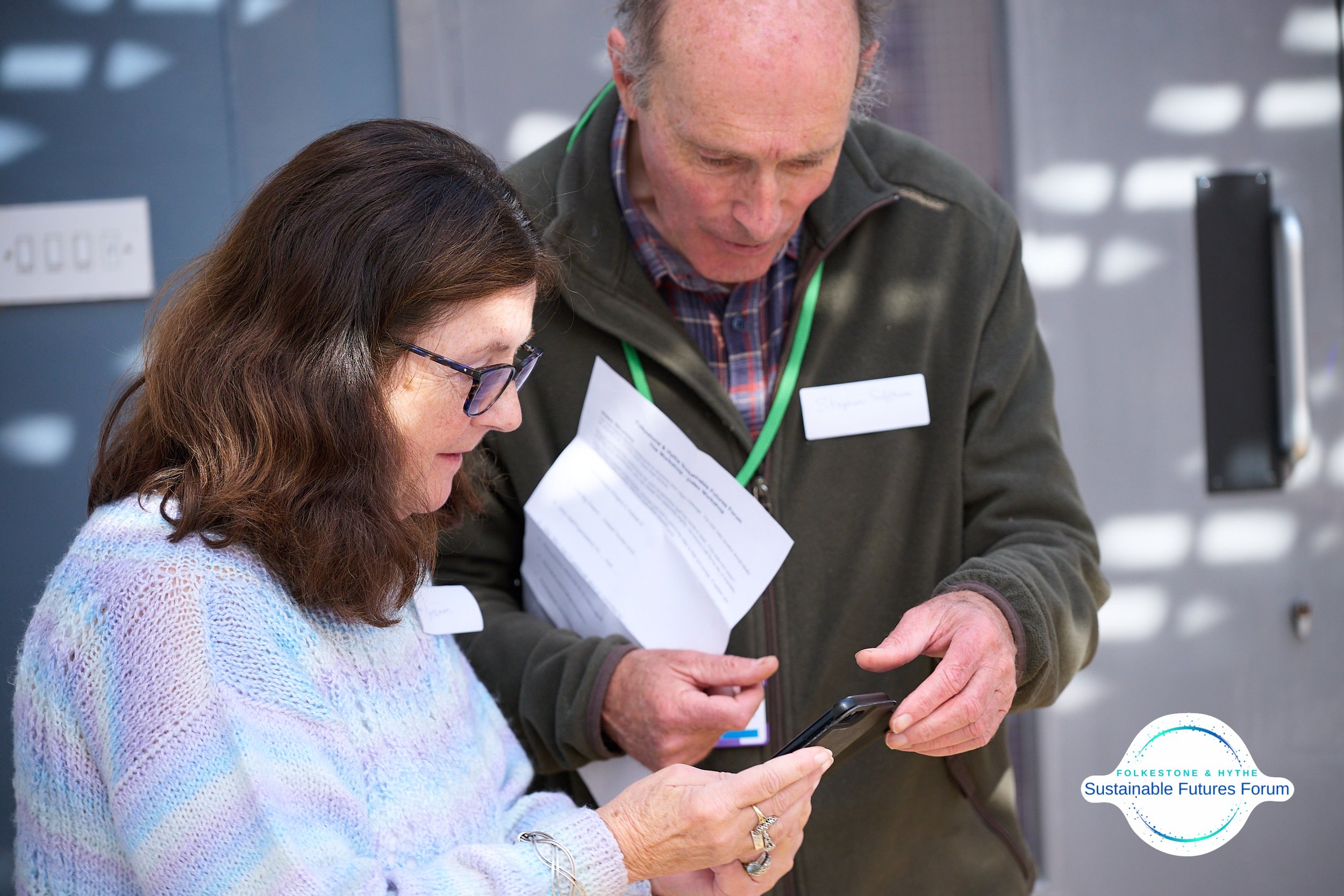 A woman and man looking at a smartphone together, with the woman holding the phone and the man pointing at it. The woman is wearing glasses and a pastel-colored sweater, while the man is dressed in a dark jacket with a name tag and holding papers. Th