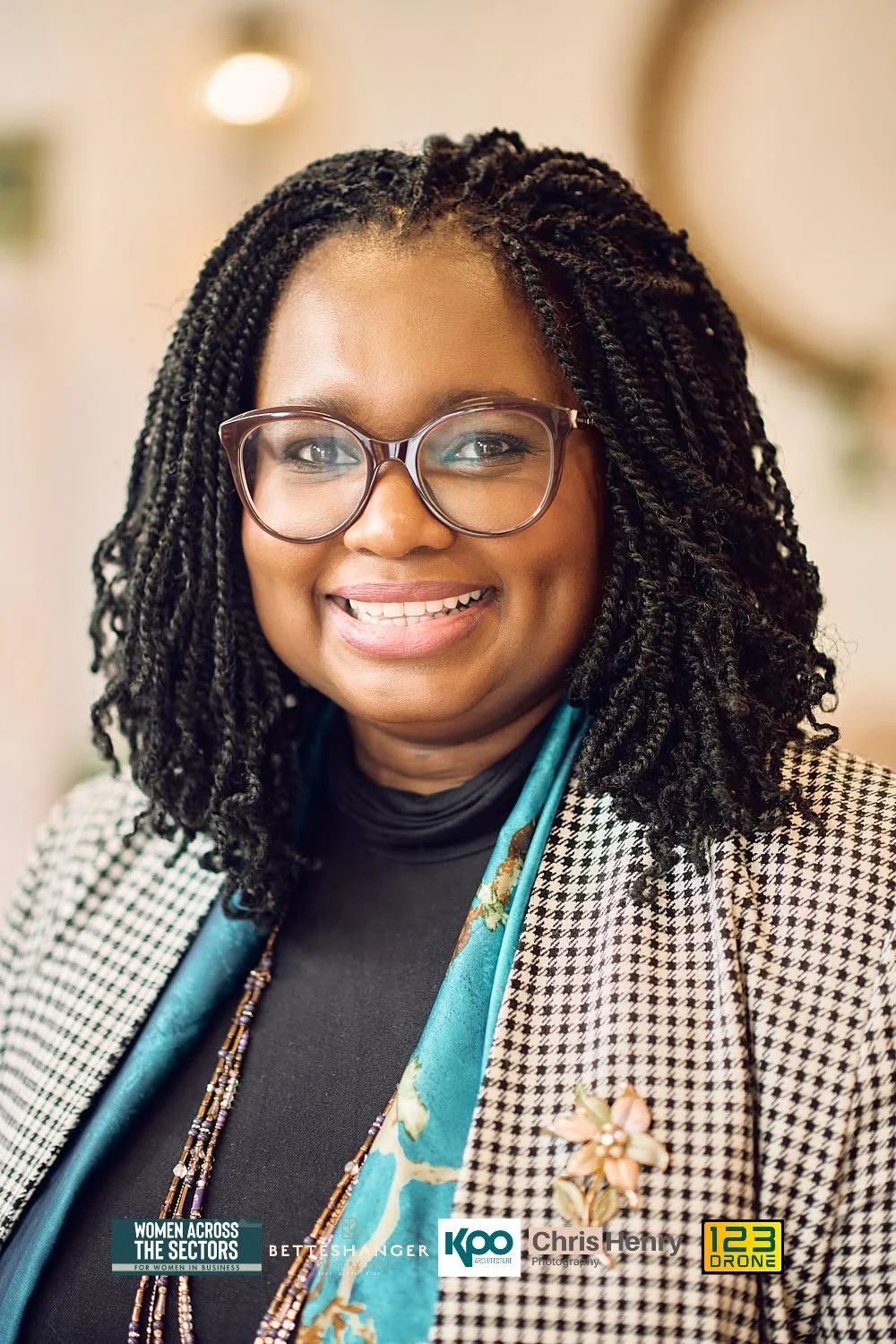A woman with glasses and styled hair smiling in an indoor setting, wearing earrings and a floral brooch, with a checkered blazer and a teal scarf.
