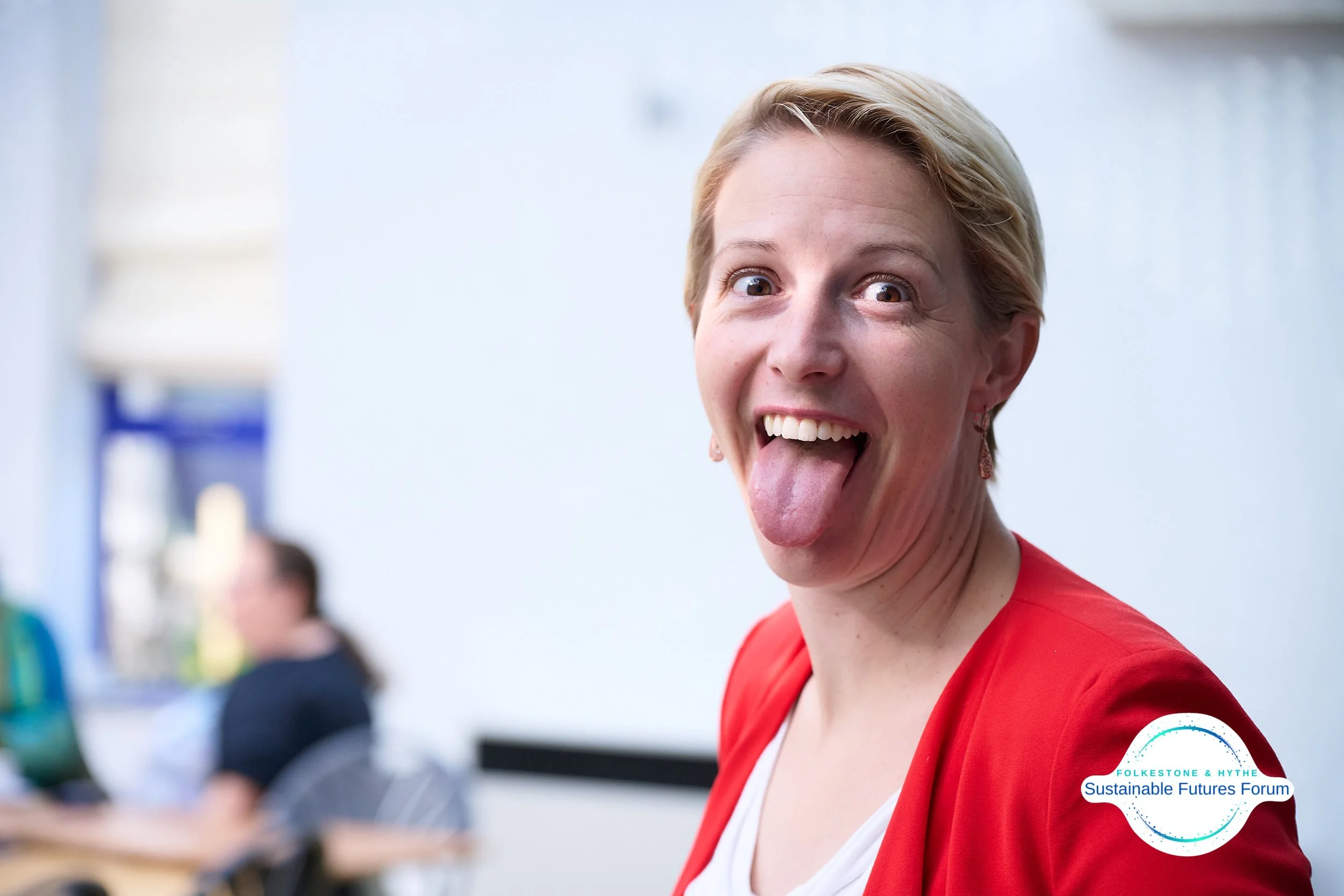 A woman sticking out her tongue and smiling, wearing a red blazer and earrings, in a classroom or conference room setting.