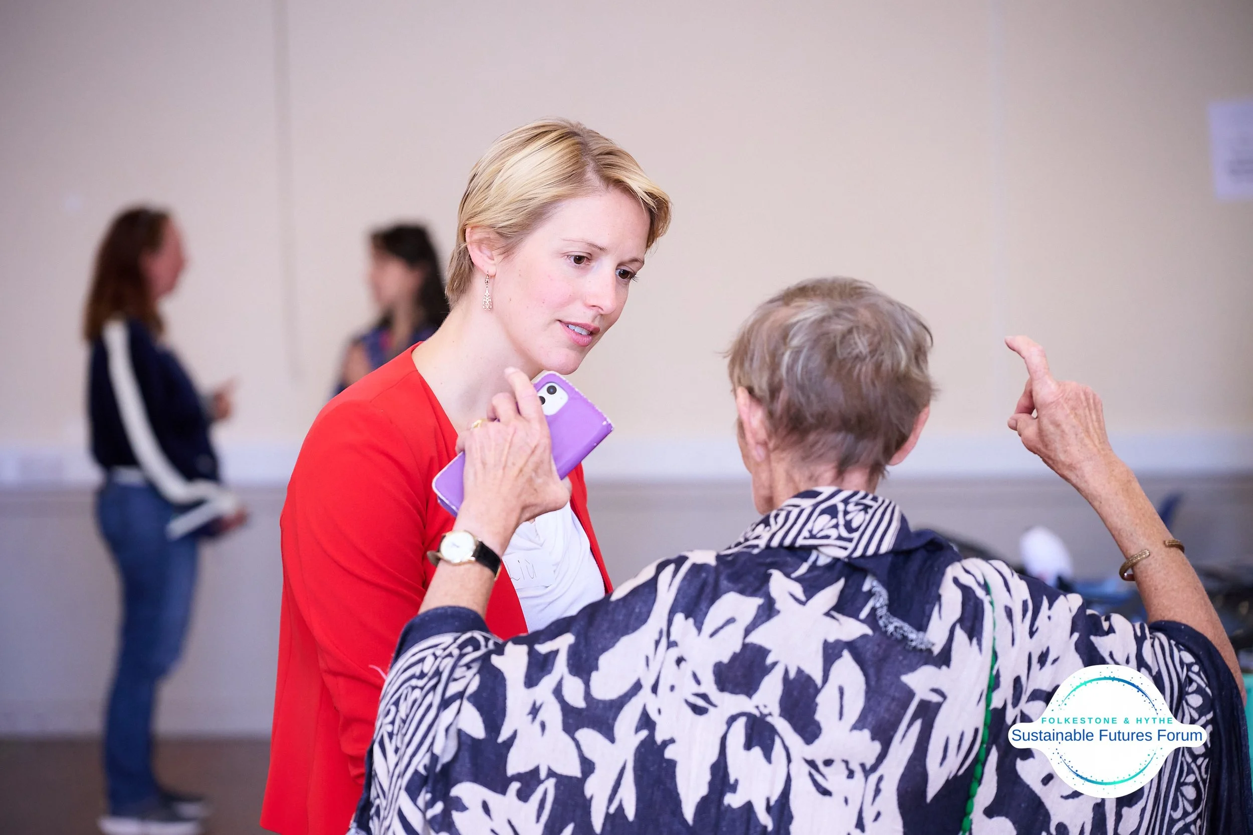 A woman in a red jacket and an elderly woman in a black and white patterned shirt are having a conversation at a community event. The elderly woman is gesturing with her hand while the woman in red holds a purple phone and listens attentively. In the
