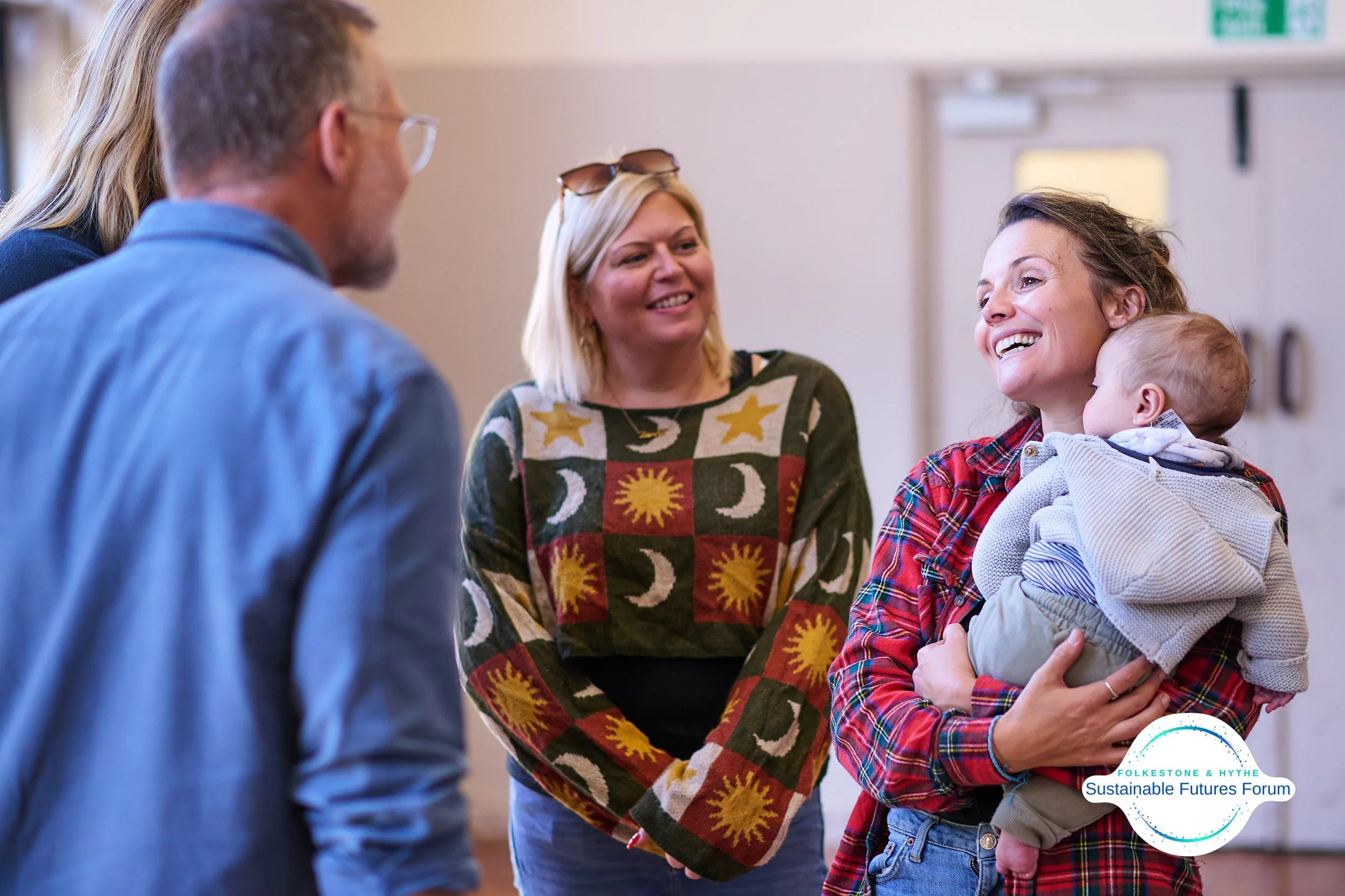 A group of four people, including a woman holding a baby, smiling and talking at an indoor forum. The woman with the baby is wearing a red plaid shirt, and the baby is in a gray hoodie. The other two adults are partially visible, one with glasses and
