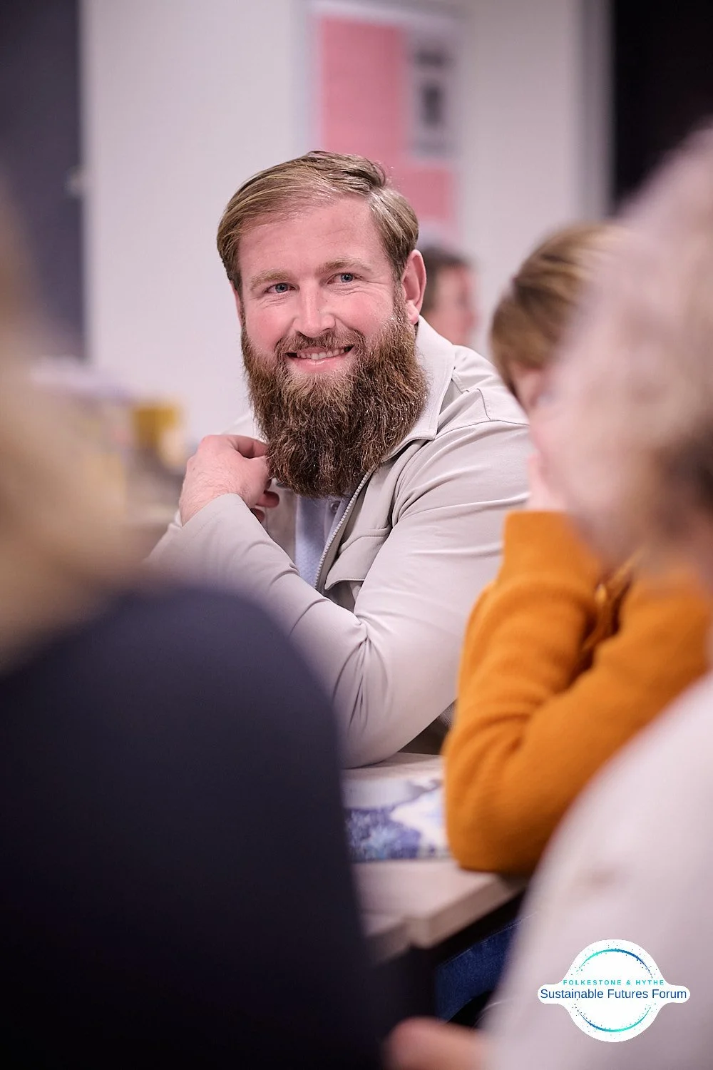 A man with a full beard smiling and looking to the side at a table during a meeting or discussion. Other people are partially visible around him. There is a logo in the bottom right corner for the Folketone & Aytche Sustainable Futures Forum.