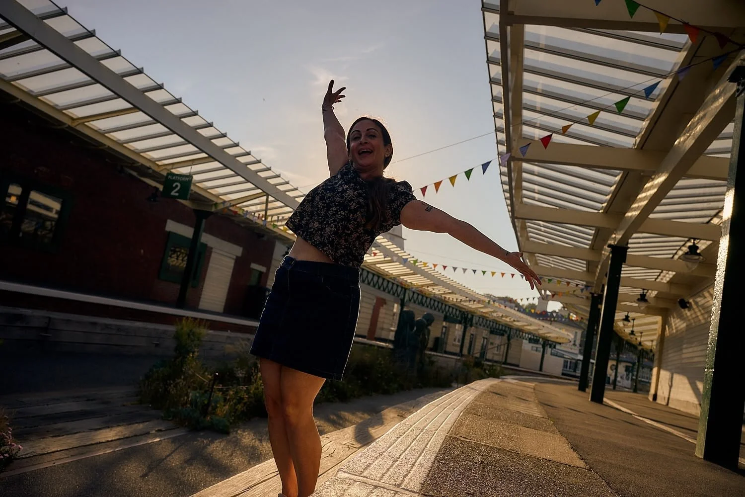 A woman dancing joyfully outdoors on a street decorated with colorful triangular flags during sunset.