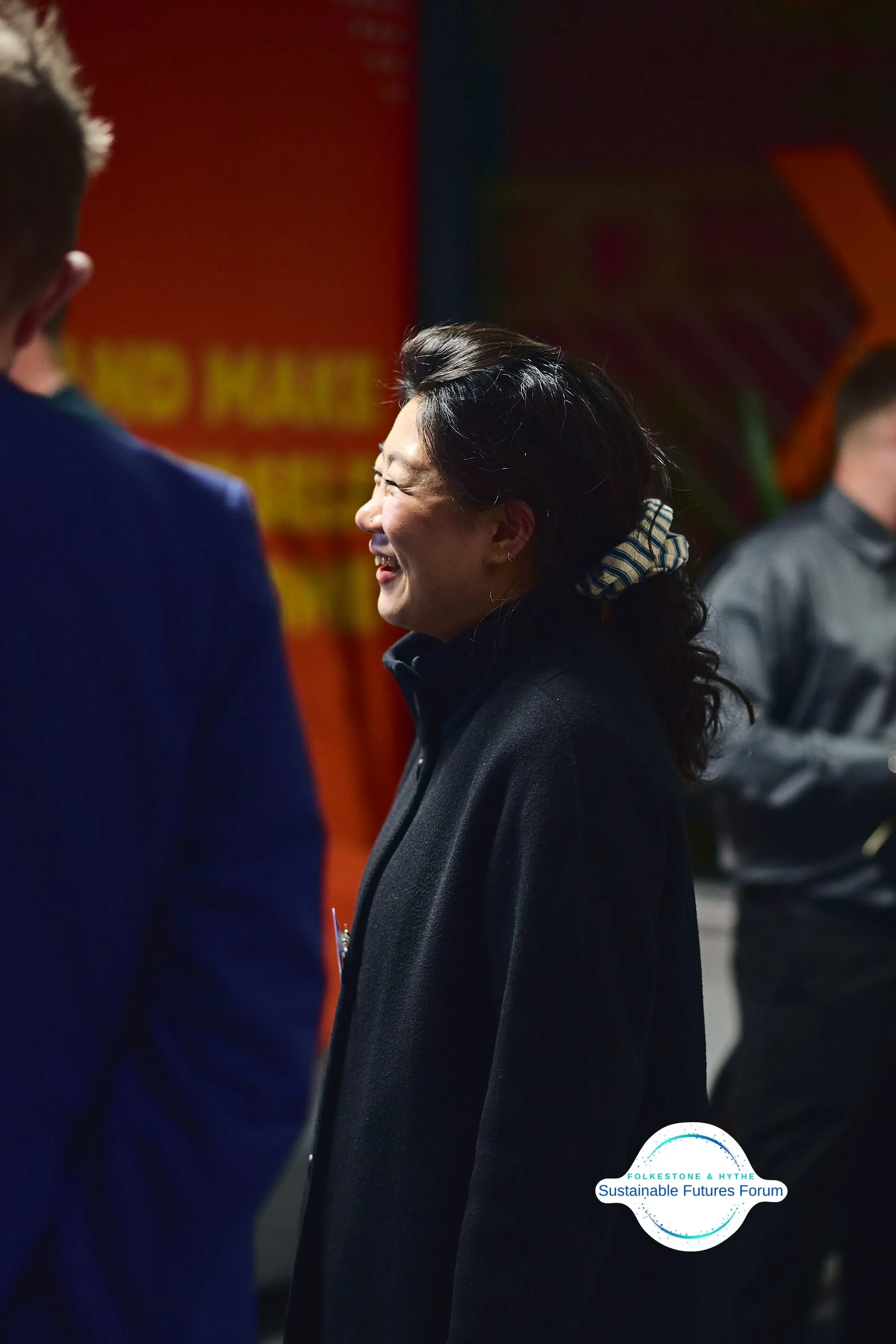 A woman with dark hair, smiling and laughing, wearing a black coat, at a forum event with a logo for Folkesone & Hythe Sustainable Futures Forum visible in the bottom right corner.