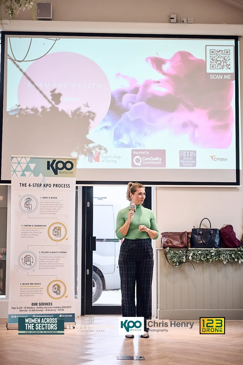 A woman in a green top and checkered pants standing in front of a presentation screen, holding a microphone, with bags and a floral decoration on a table behind her. The screen displays information about hormone health, a QR code, and logos of health