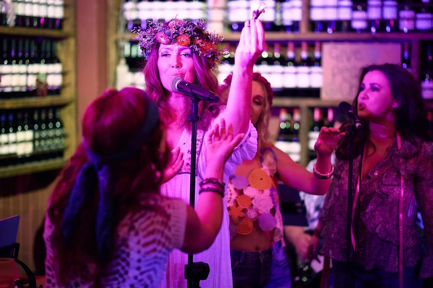 Women singing and singing karaoke in a bar, one woman wearing a flower crown and a white dress.