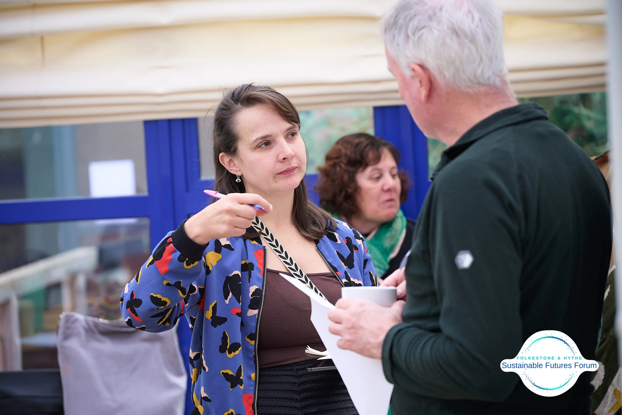 Two women and a man engaged in a conversation at an indoor event. The woman in the foreground is holding a pen and wearing a colorful butterfly-patterned jacket, listening to the man. The woman in the background has curly hair and is wearing a green 