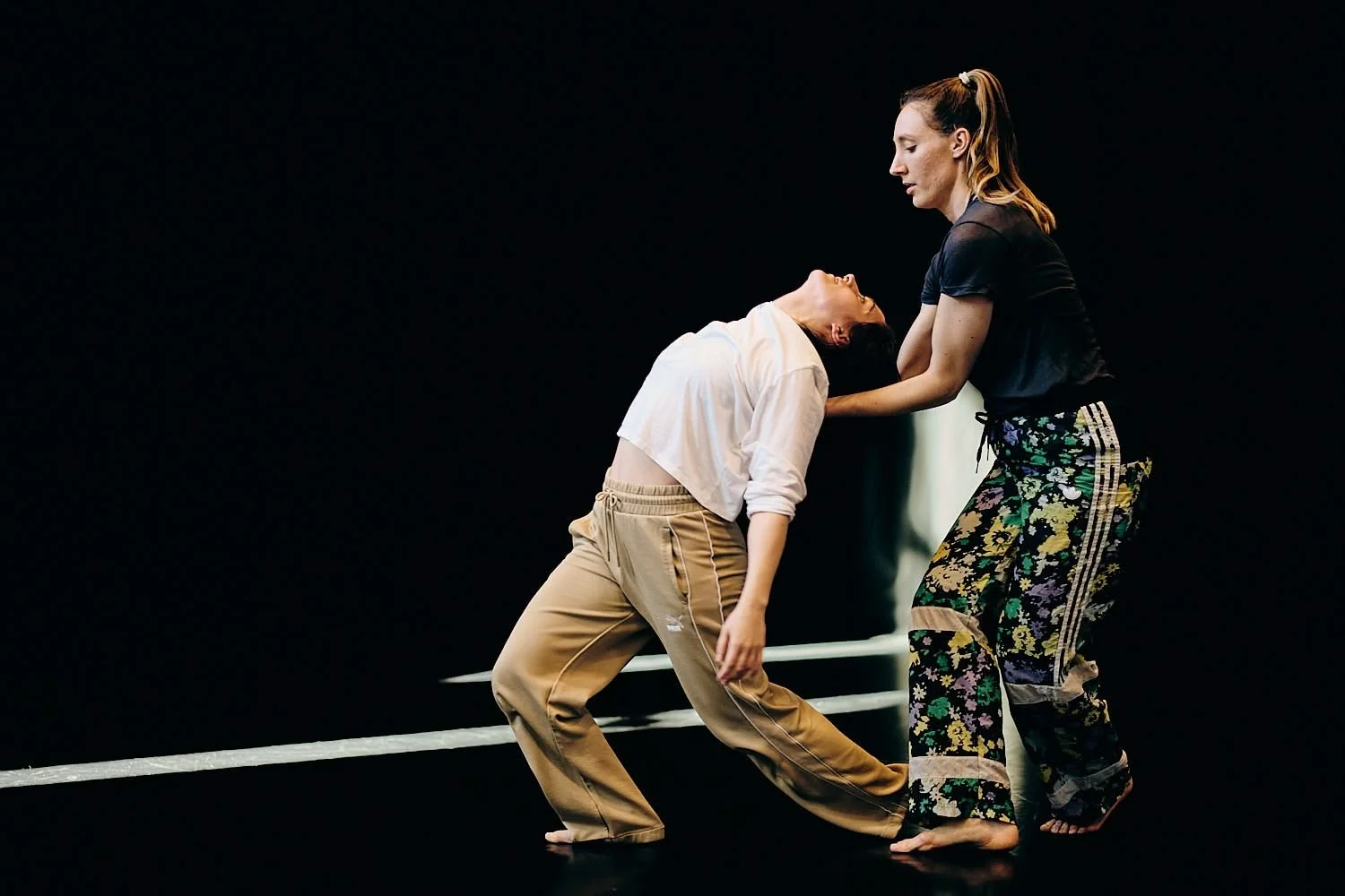 Two women practicing dance in a studio with a dark background, one is kneeling and the other standing, holding her by the shoulders.