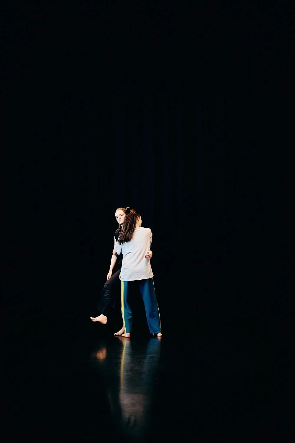 Two people hugging on a dark stage, one in a white shirt and the other in a black shirt, with a black background and reflective floor.