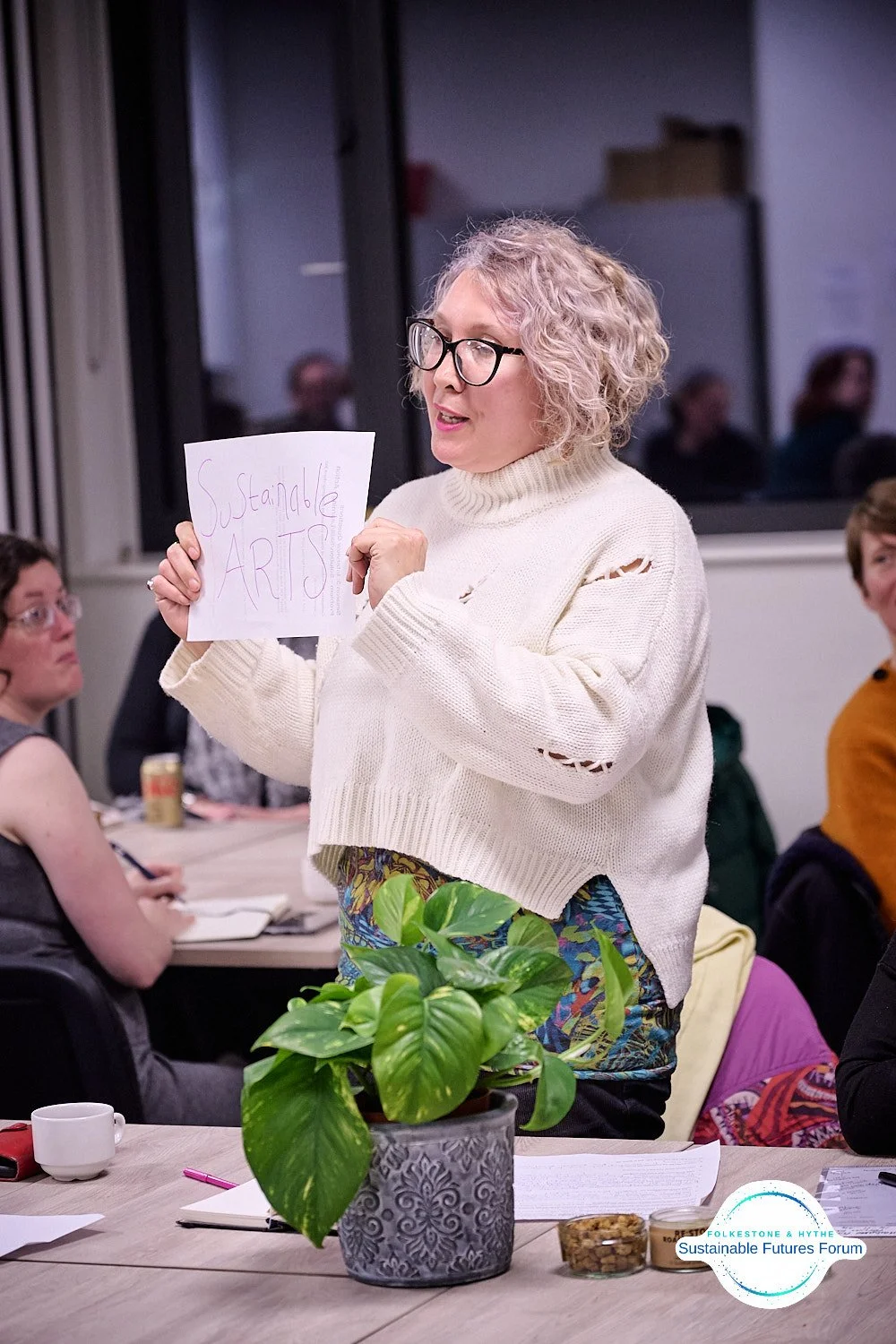 Woman with curly blonde hair and glasses holding a sign that reads 'Sustainable Arts' at a meeting or workshop, with people seated at tables and a potted plant in the foreground.