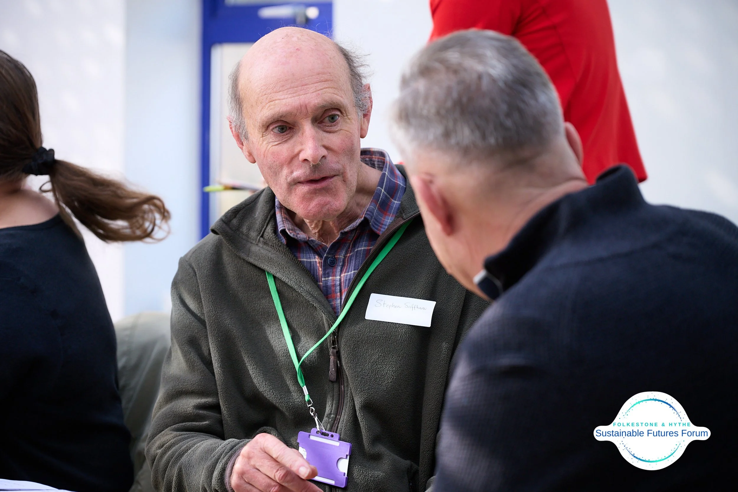 Three people in conversation at the Sustainable Futures Forum, including an older man with a name badge for Stephen Saffran, wearing a gray jacket and a plaid shirt, with blurred details of other participants.