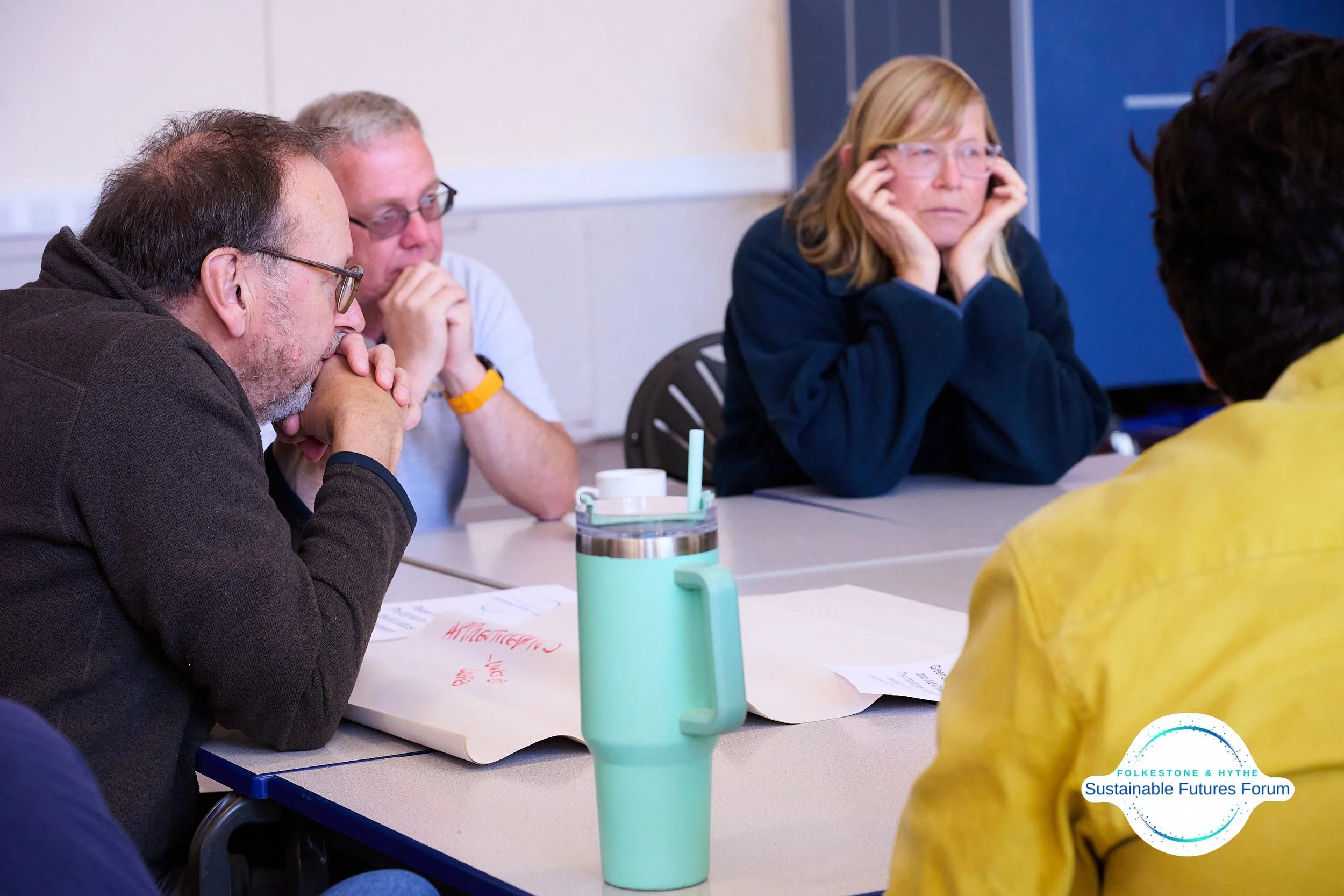 A group of five adults sitting at a table during a discussion or meeting, with papers and a water tumbler in the foreground.