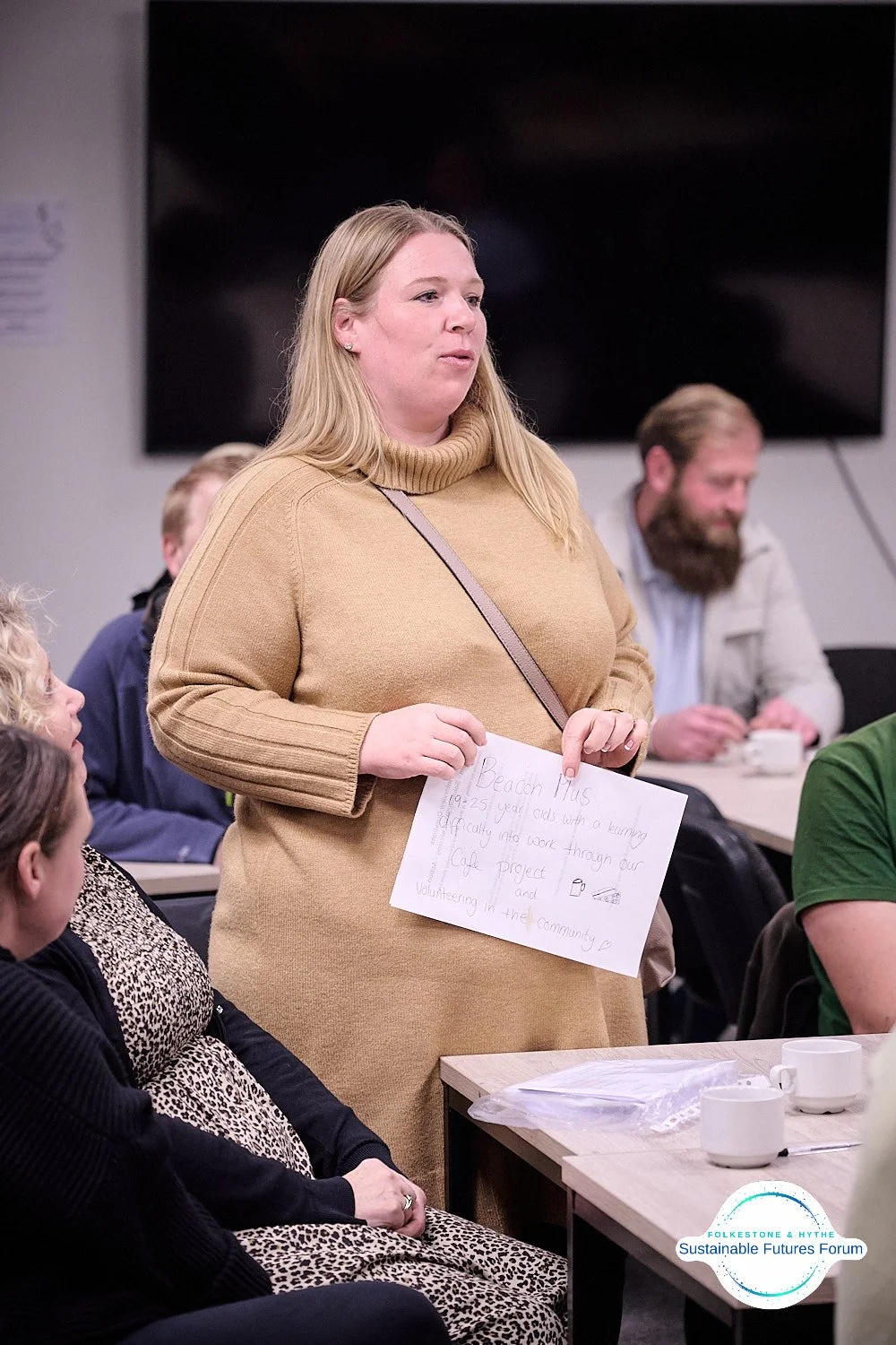 A woman in a tan turtleneck sweater holds a handwritten note at a community meeting, with other attendees seated around her.