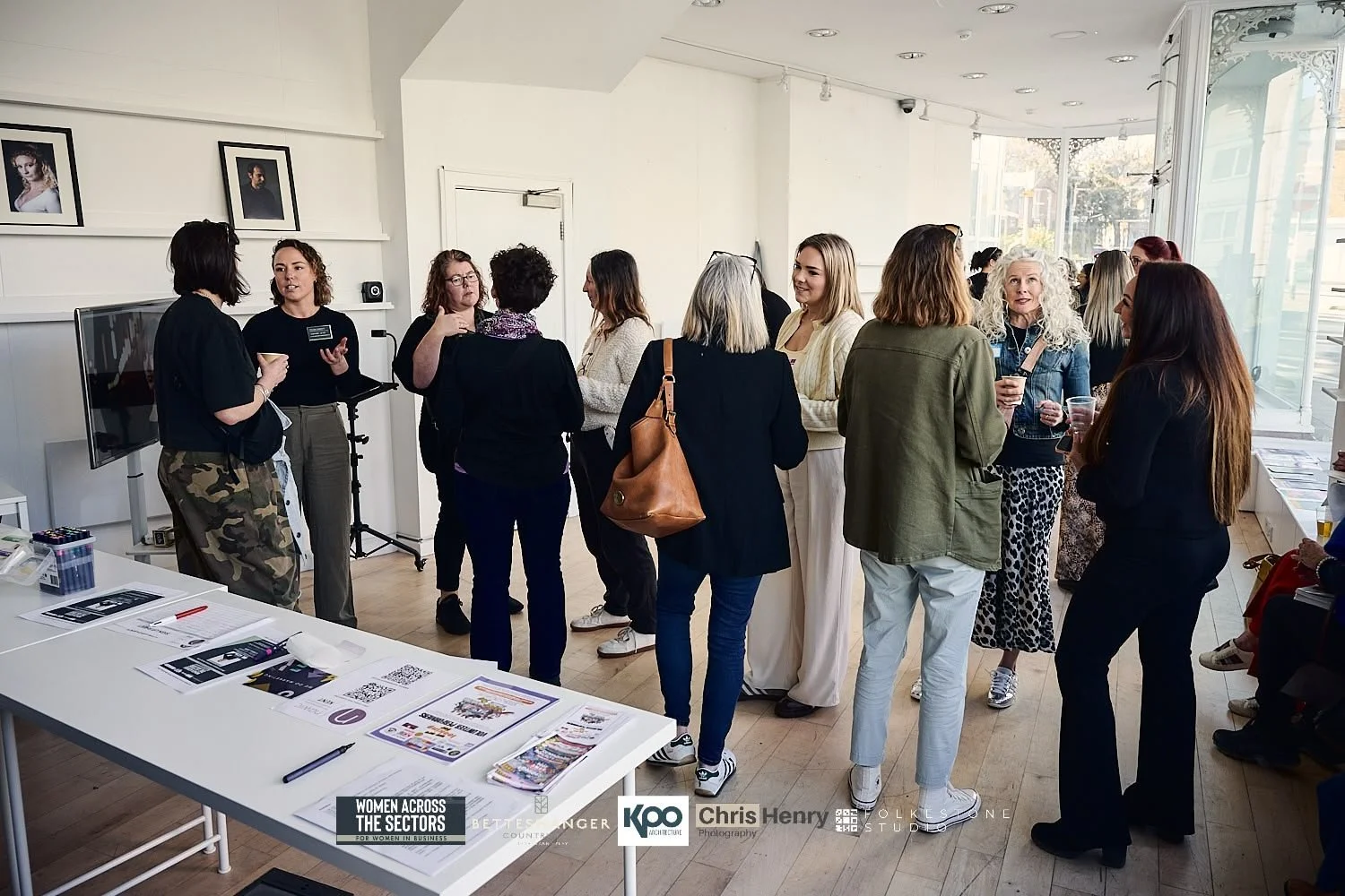 Women attending a networking or social event in a bright, modern room with white walls and large windows. Some women are conversing in groups, holding drinks, and engaging in discussions, with informational materials on a table in the foreground.
