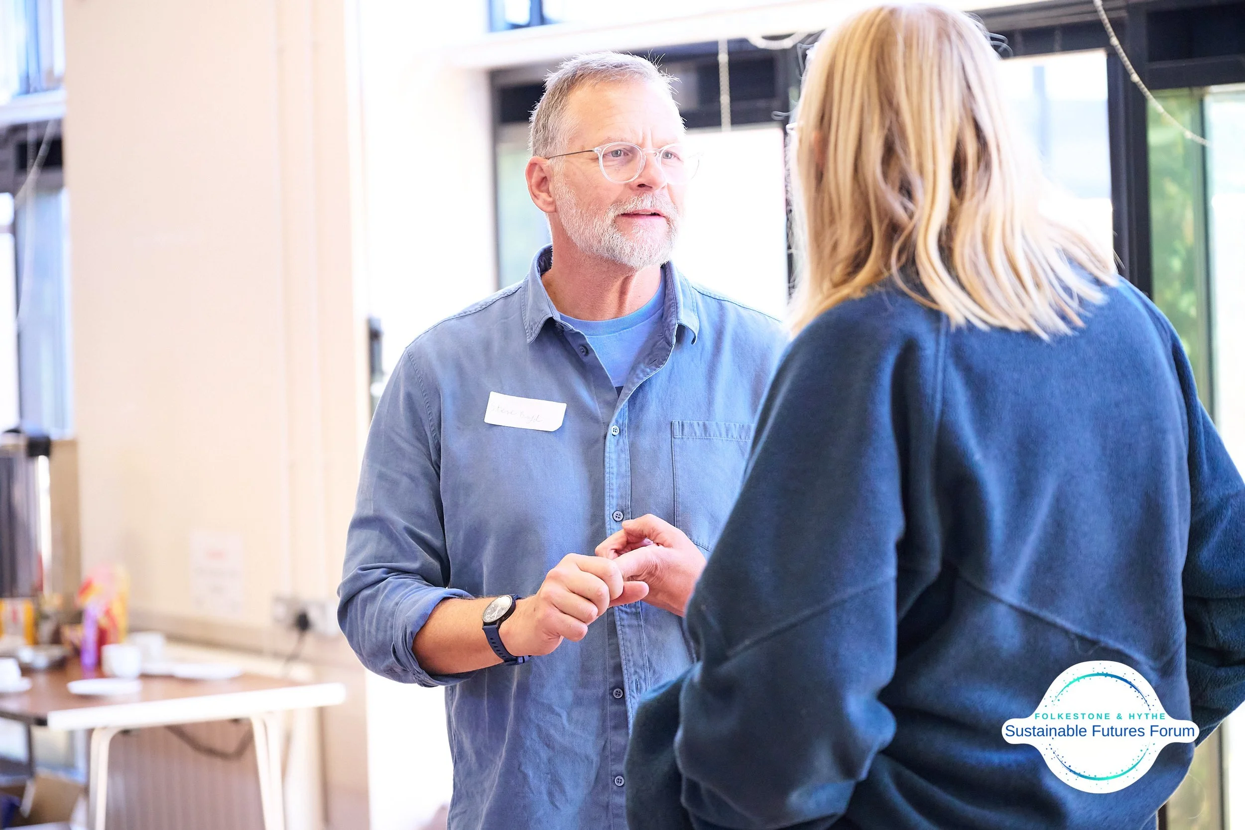 A man and a woman are having a conversation indoors, with the man holding the woman's hand. The man has glasses, a gray beard, and is wearing a blue shirt with a name tag, and the woman has blonde hair and is wearing a navy sweater. There is a logo i