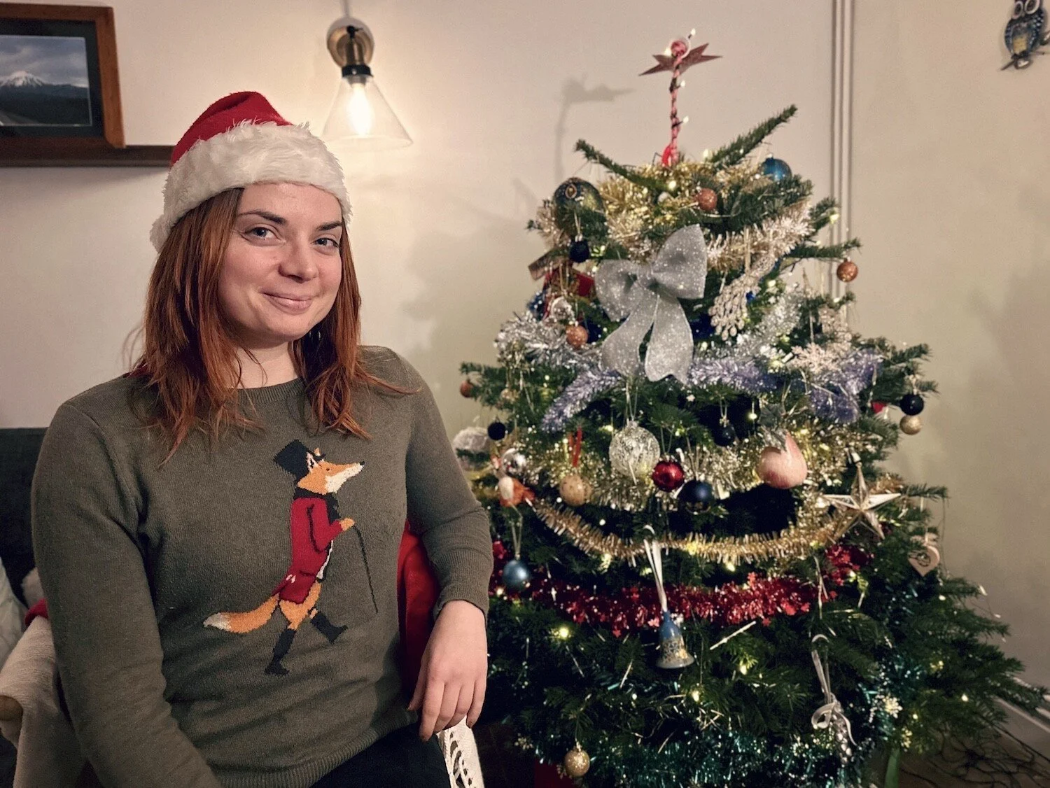 Woman in green jumper and Santa hat next to Christmas tree