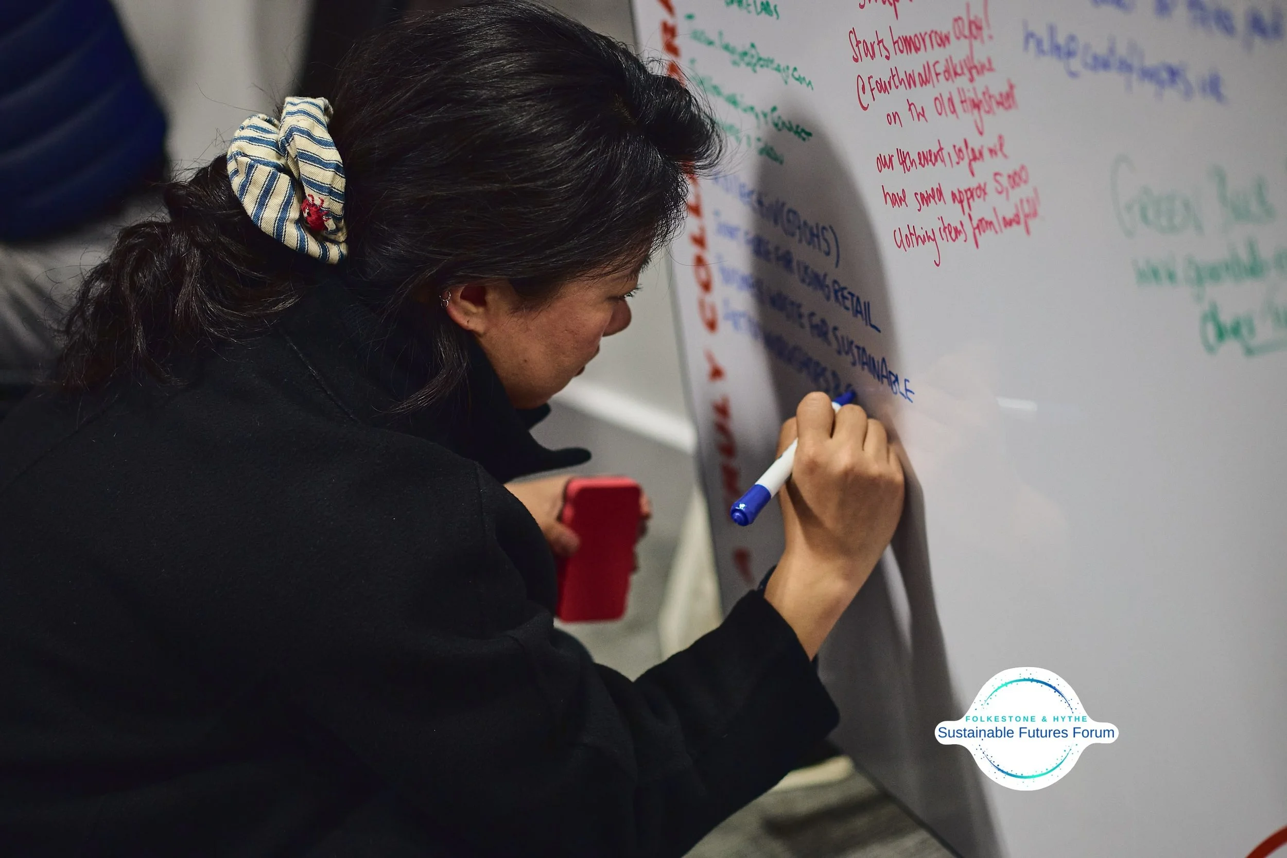 A woman with dark hair, wearing a striped scrunchie, writing on a large whiteboard with a blue marker, while holding a red smartphone in her left hand. The whiteboard contains colorful handwritten notes and has a "Sustainable Futures Forum" logo at t