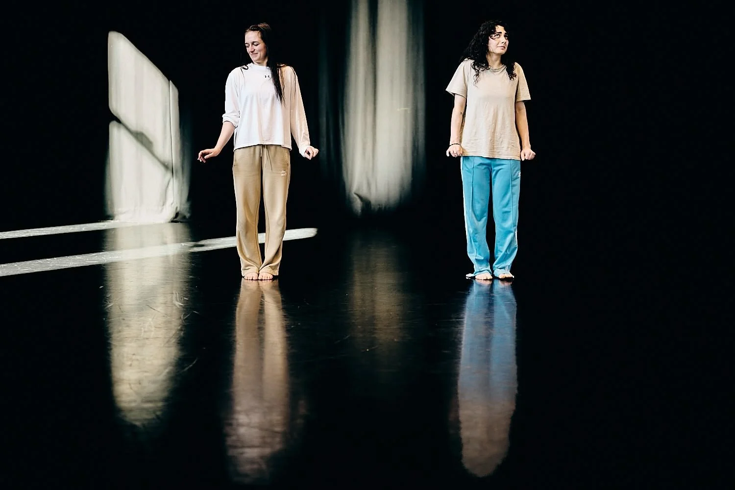 Two women standing on a black stage with reflections, wearing comfortable clothes, one smiling and the other looking serious.
