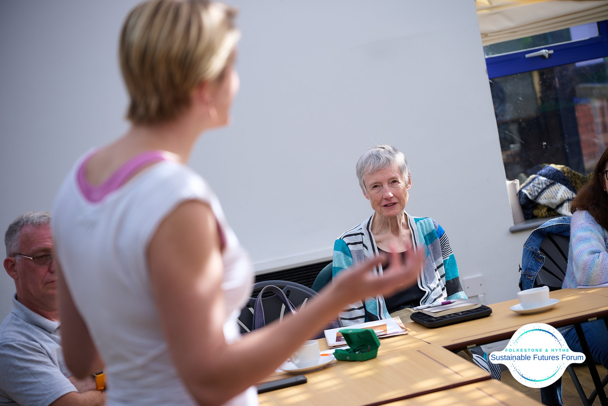 A woman with short blonde hair is speaking and gesturing with her hand, while an elderly woman with short gray hair listens attentively at a table during a meeting or discussion at the Folkestone & Hythe Sustainable Futures Forum.
