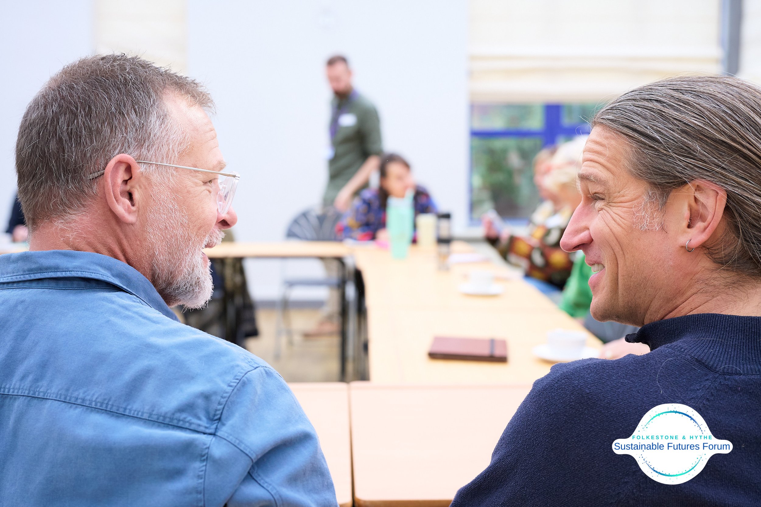Two men are smiling and talking in a meeting room with a table, other people are in the background. The logo in the bottom right corner reads 'Folkestone & Hythe Sustainable Futures Forum'.