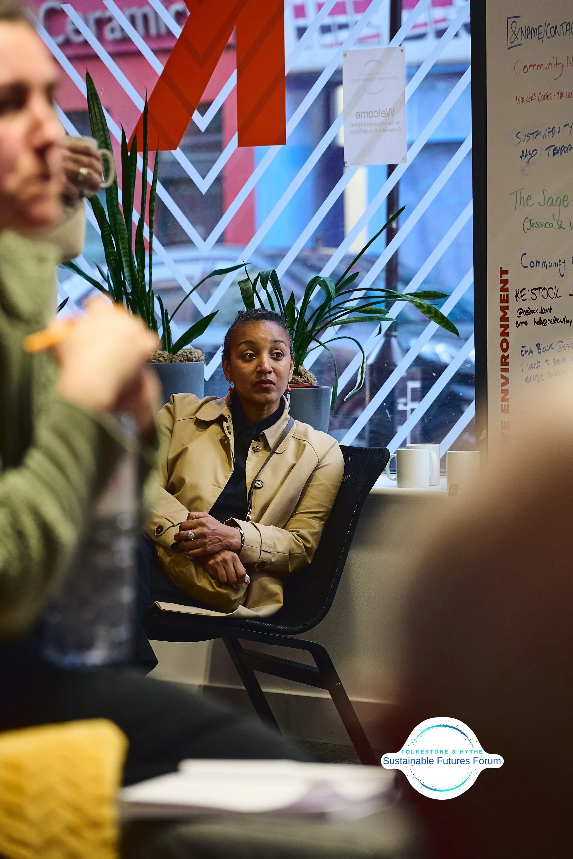Woman sitting and listening at a meeting with a whiteboard and plants in the background.