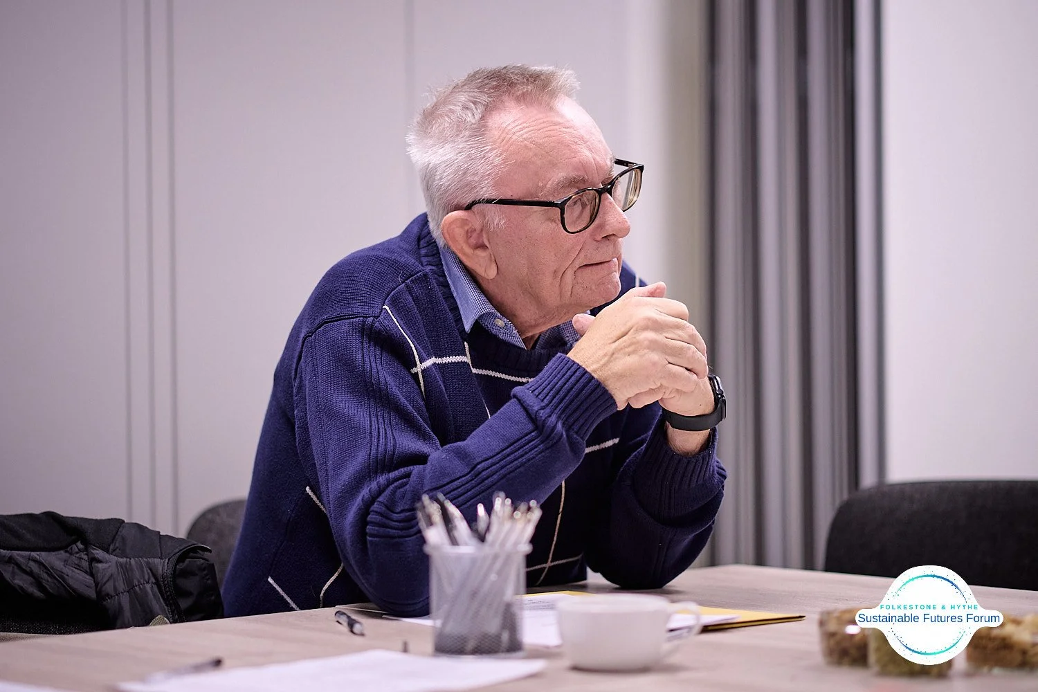 An elderly man with glasses and gray hair, wearing a blue sweater with white stripes over a collared shirt, sits at a conference table with his hands clasped in front of his face, appearing thoughtful. There are papers, a pen, a cup, and a container 
