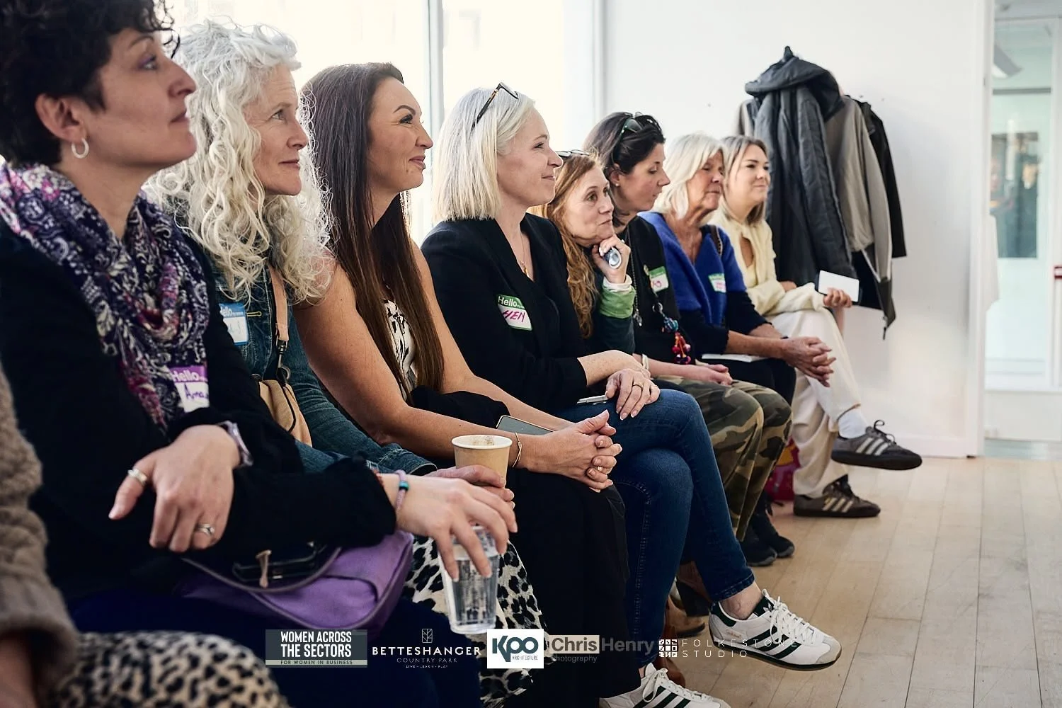 Group of women sitting in a row at an indoor event, some holding cups and glasses, listening attentively.