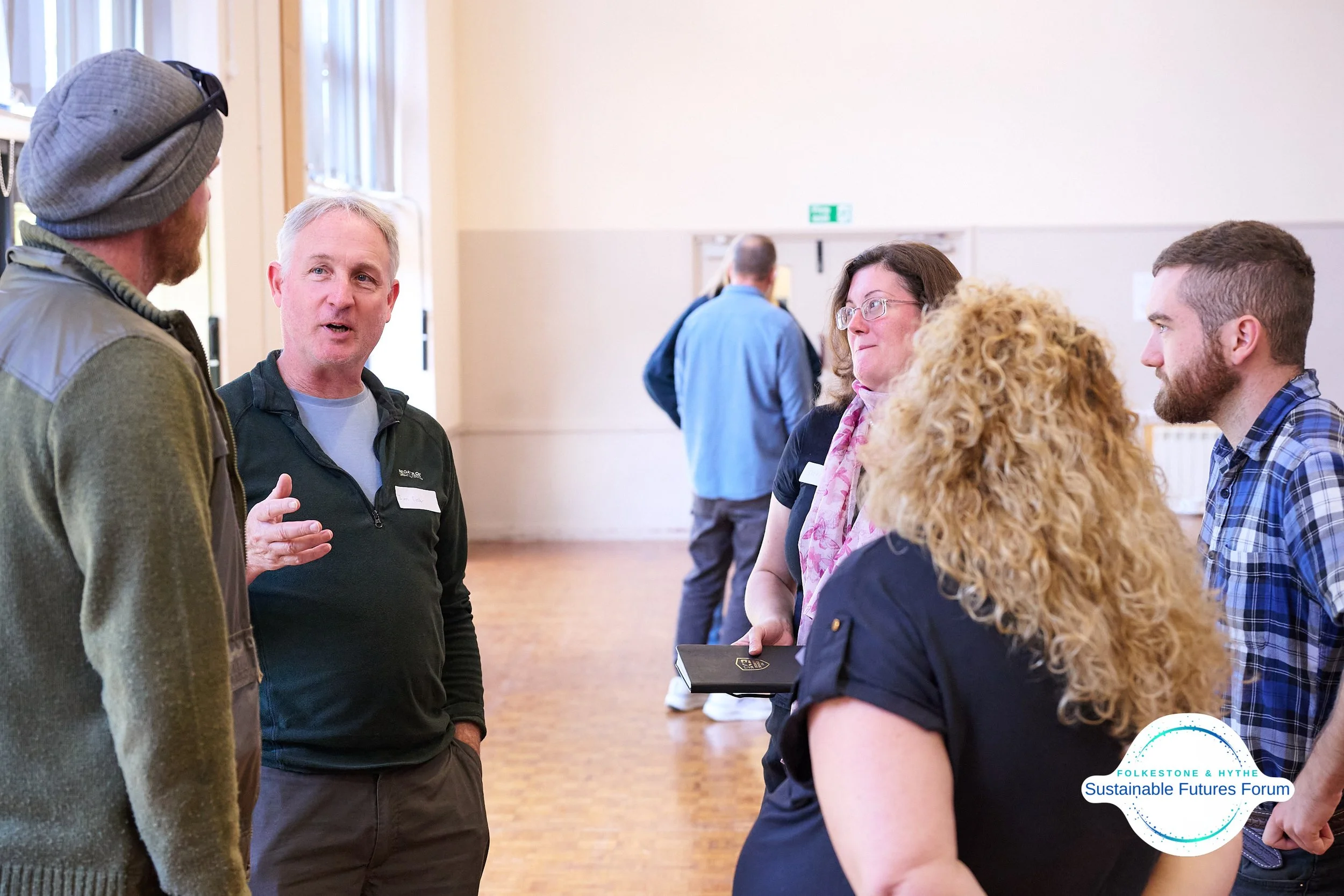 Group of people engaged in conversation at a community event, with a logo for Folksstone & Hythe Sustainable Futures Forum in the corner.
