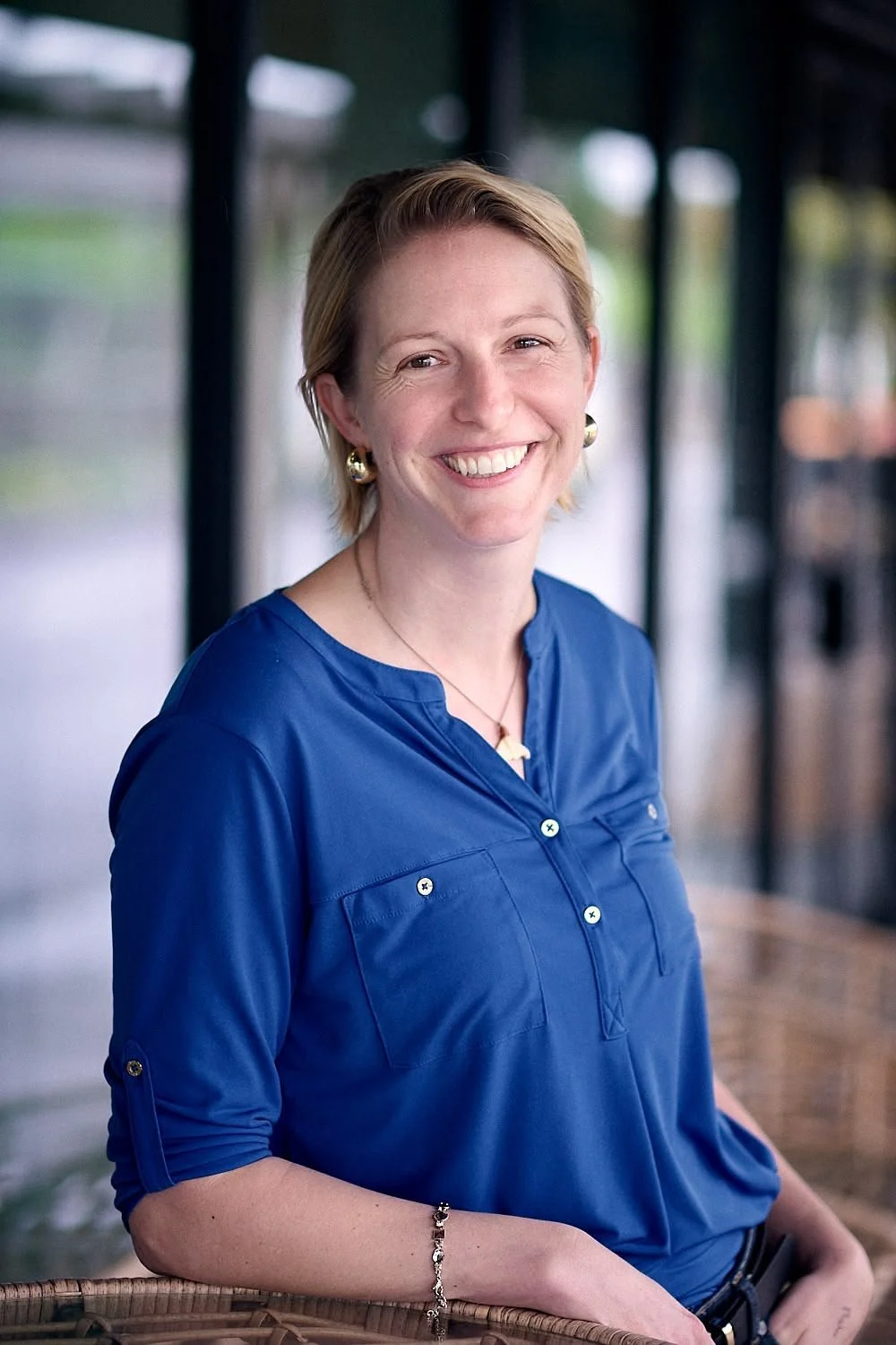 A woman with short blonde hair smiling, wearing gold earrings, a necklace, a blue blouse with rolled-up sleeves, sitting outdoors in a covered area with blurred background.