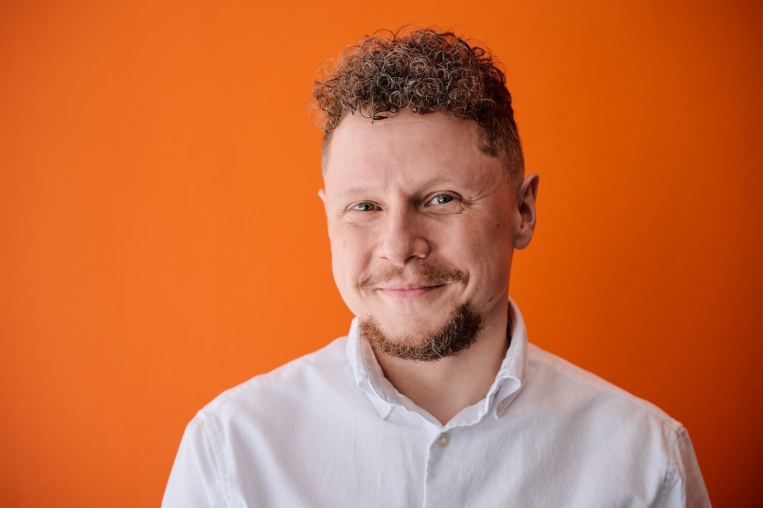 Portrait of a man with curly hair, a goatee, wearing a white shirt, smiling against an orange background.