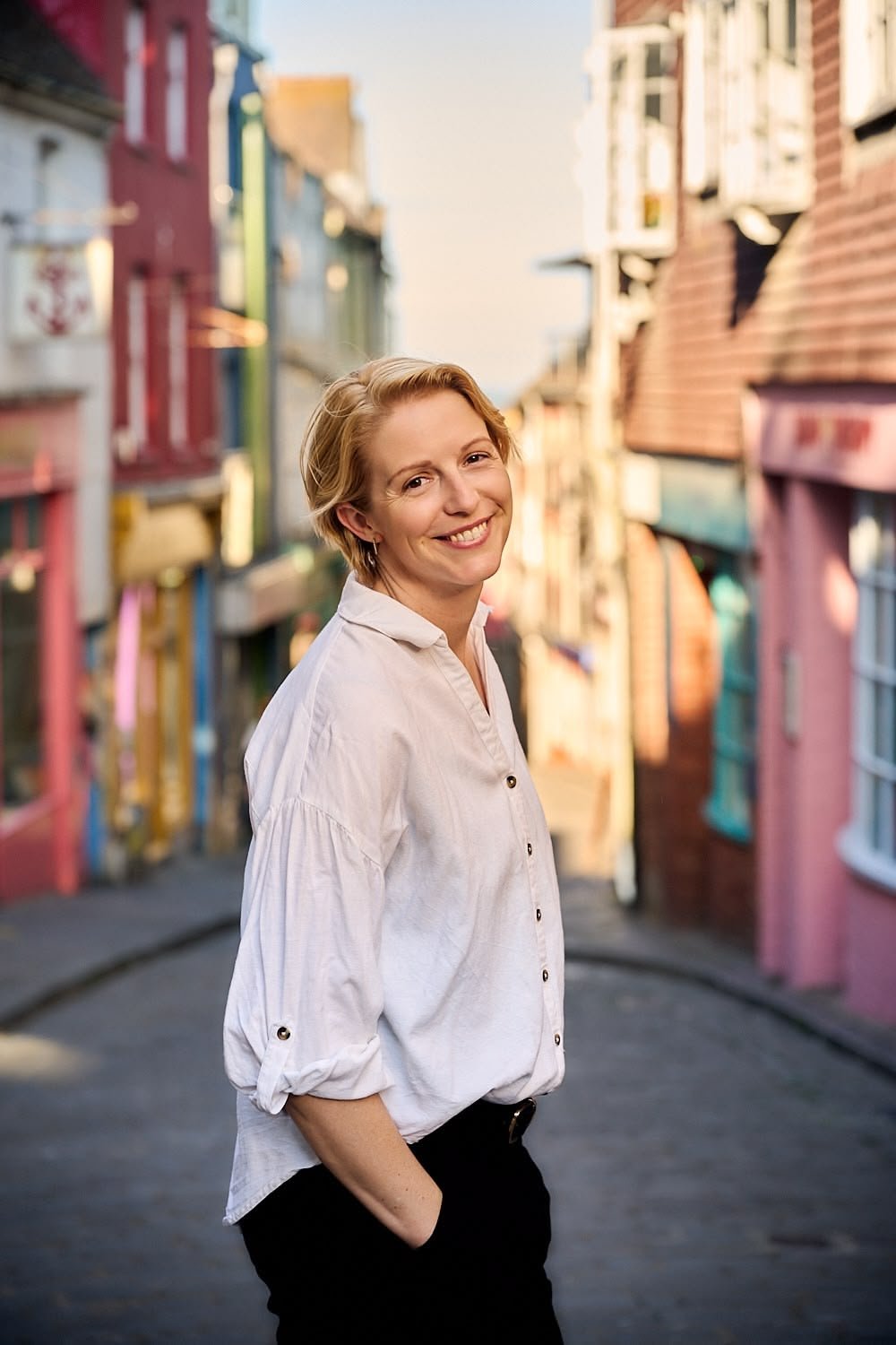Smiling woman with short blonde hair in a white shirt standing in a colorful alleyway with buildings painted in pastel colors.