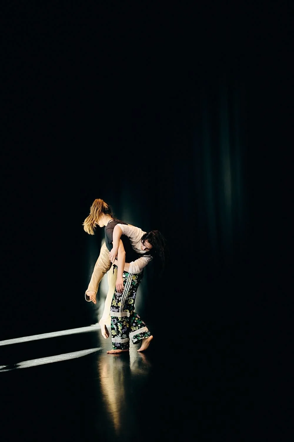 Two dancers perform on a dark stage with minimal lighting, one lifting the other in a contemporary dance pose.