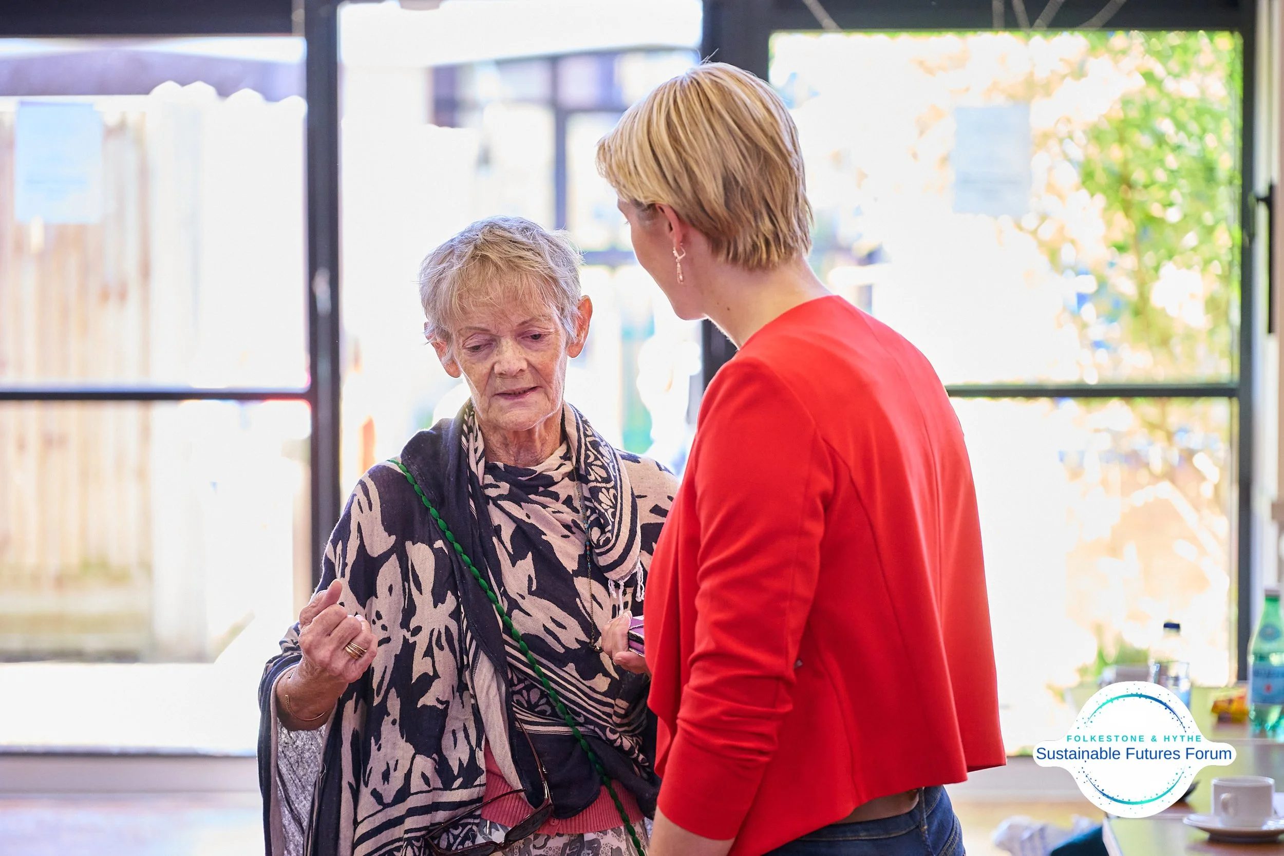 A woman in a red jacket talking to an elderly woman with short gray hair inside a well-lit room with large windows, a small table with a cup and bottle, and a woodland view outside.