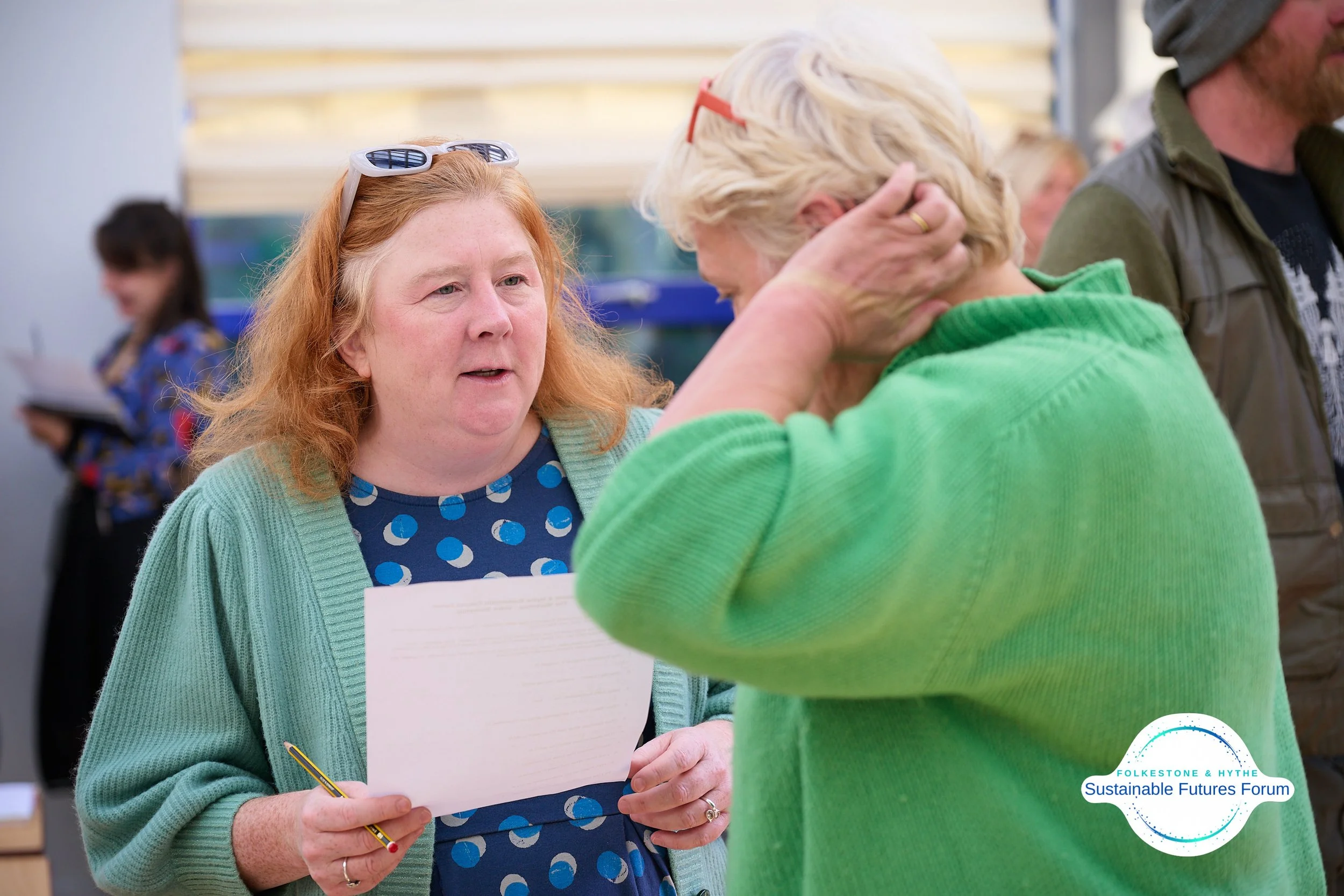 Two women are engaged in a conversation at a forum, with one woman holding a piece of paper and a pen. The woman on the left has red hair, is wearing a aqua cardigan and a blue dress with a circular pattern. The woman on the right has blonde hair, is