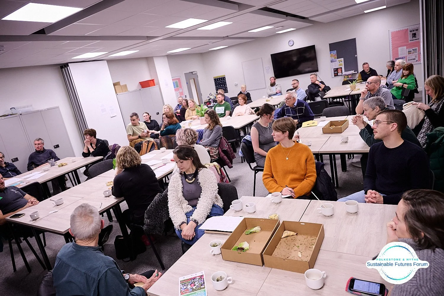 A group of people attending a meeting or workshop in a conference room, sitting mostly at tables with paper, cups, and some leftover food, while listening to a speaker or presentation.