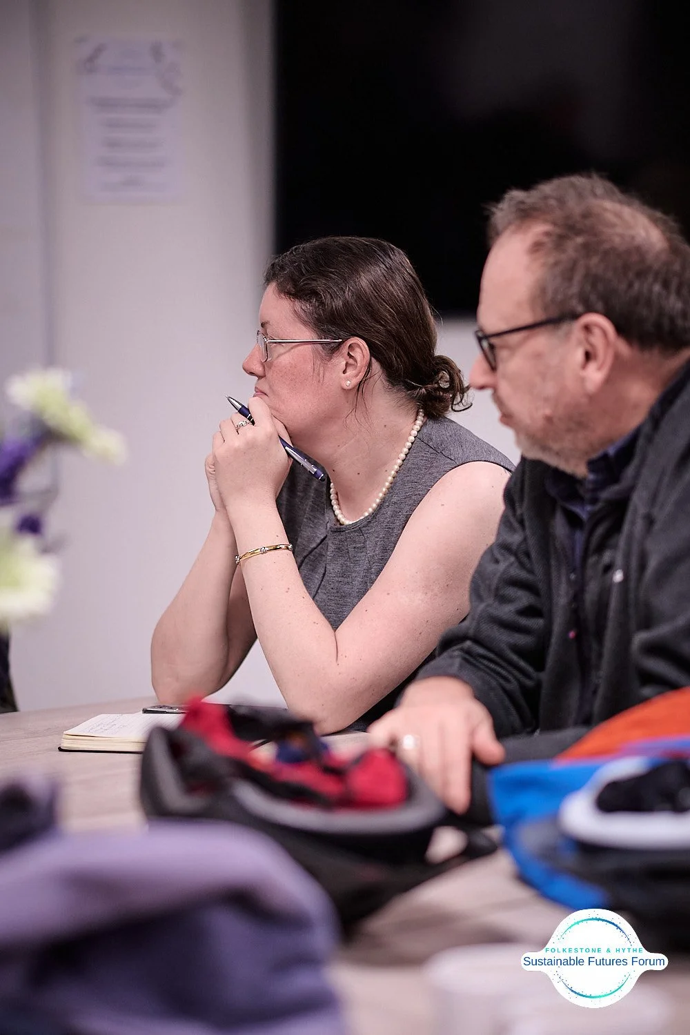 A woman wearing glasses, a gray sleeveless top, a pearl necklace, and a gold bracelet sitting at a table with her hand resting on her chin, holding a pen, during a meeting at the Sustainable Futures Forum. A man next to her, also wearing glasses, is 