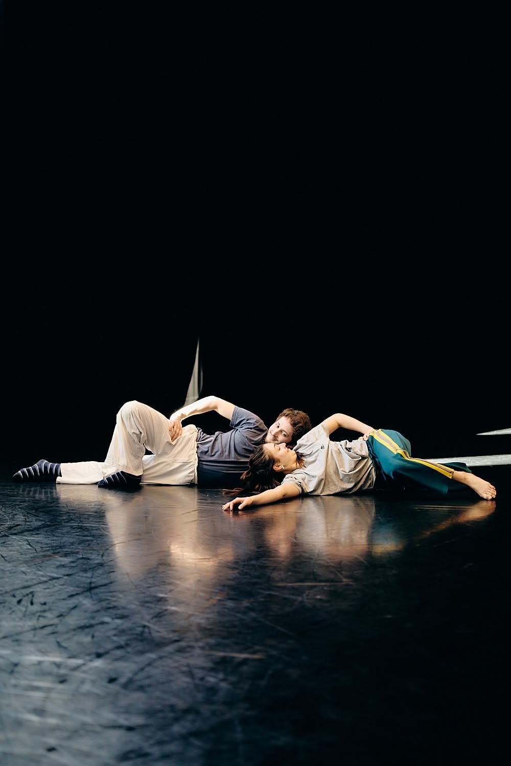 Two performers lie on a dark stage, embracing each other with contrasting postures, one with a man on his side and a woman on her back, in a theatrical or dance rehearsal setting.