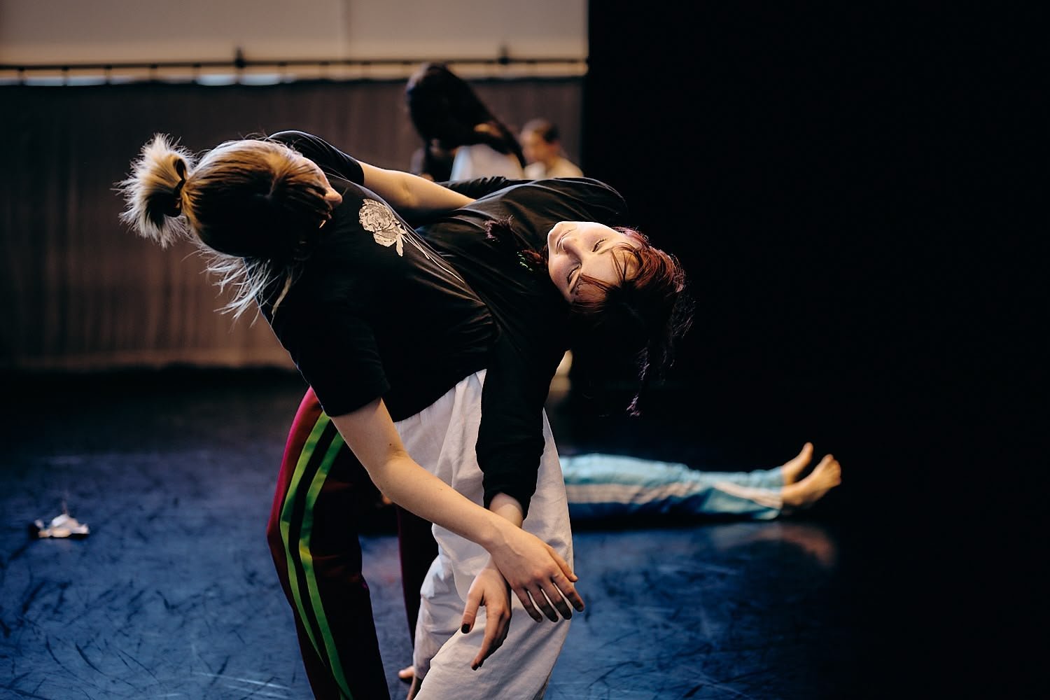 Two women practicing acro yoga or a similar physical activity on a dark floor in a dance studio, with other participants visible in the background.