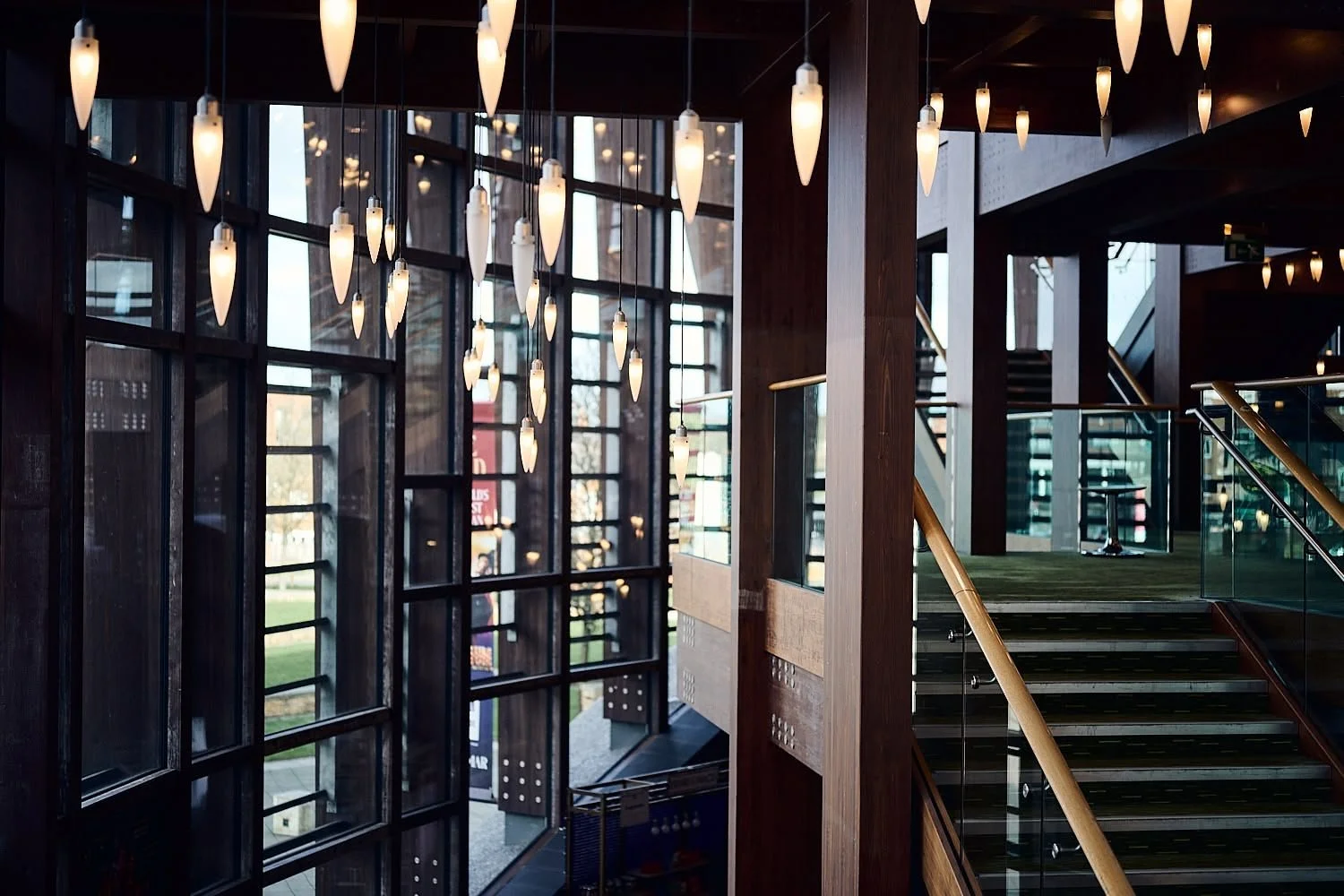 Interior of a modern building with large windows, hanging pendant lights, wooden and glass staircase, and natural light coming through the windows.