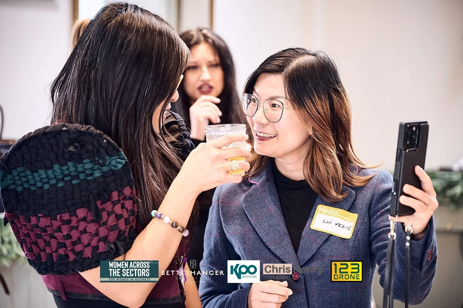 Three women at a social event, one woman is holding a glass of beverage while taking a selfie with a smartphone, another woman is in the background. The woman taking the selfie is wearing glasses and a name tag that says 'Chi Years.'