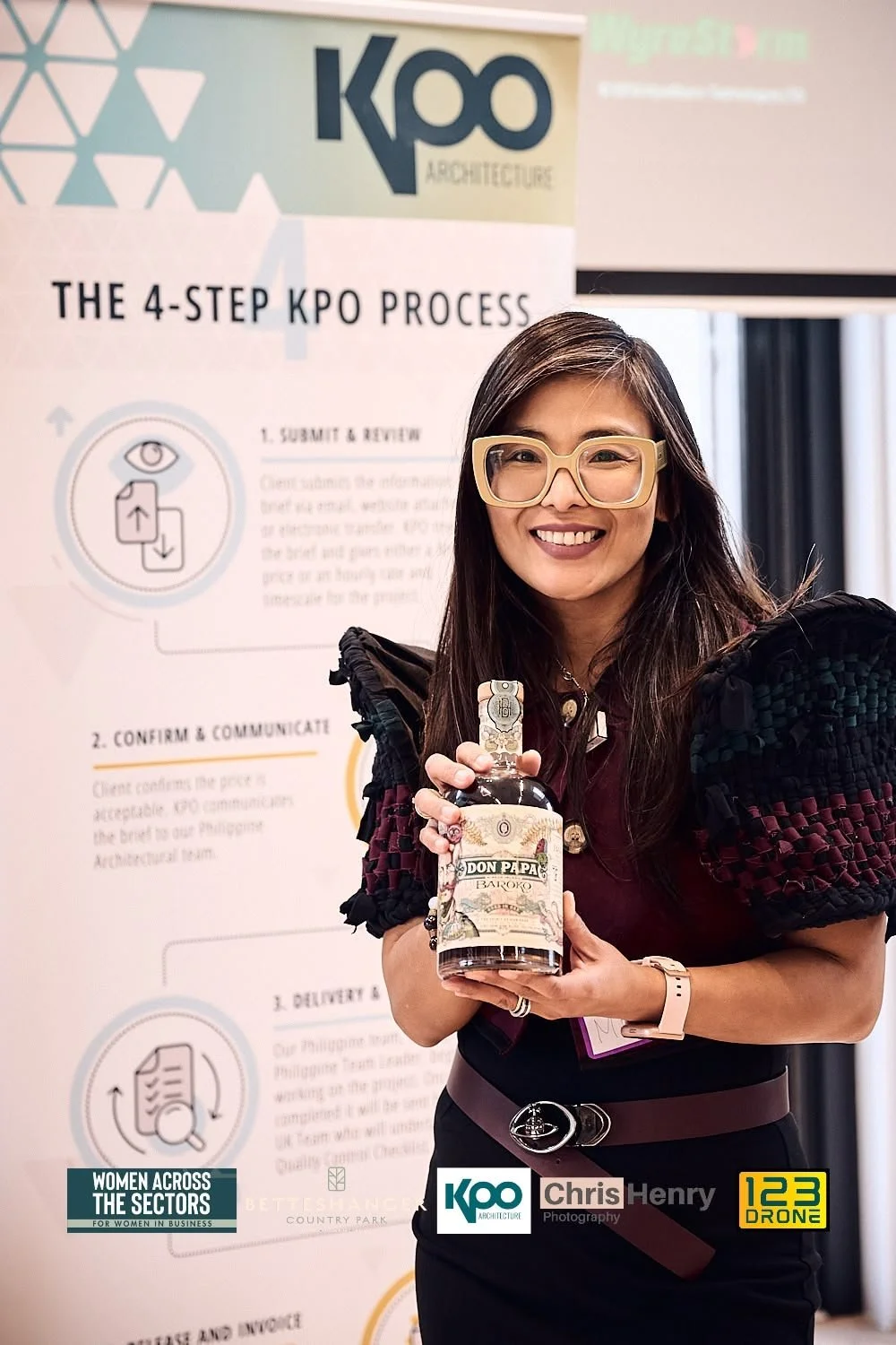 A woman with long dark hair and glasses smiling, holding a bottle of Don Papa rum, standing in front of a KPO Architecture poster about the 4-step KPO process.
