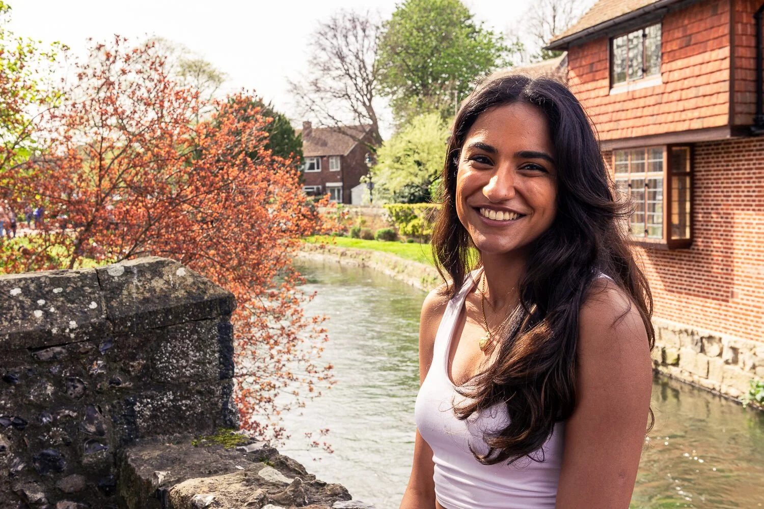 A smiling woman with long dark hair standing by a river on a sunny day, with trees and houses in the background.