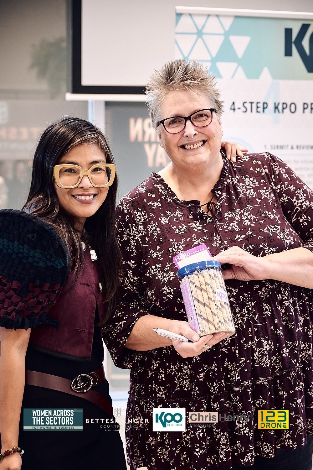 Two women smiling at a professional event, one holding a container of cookies, with promotional banners and logos in the background.